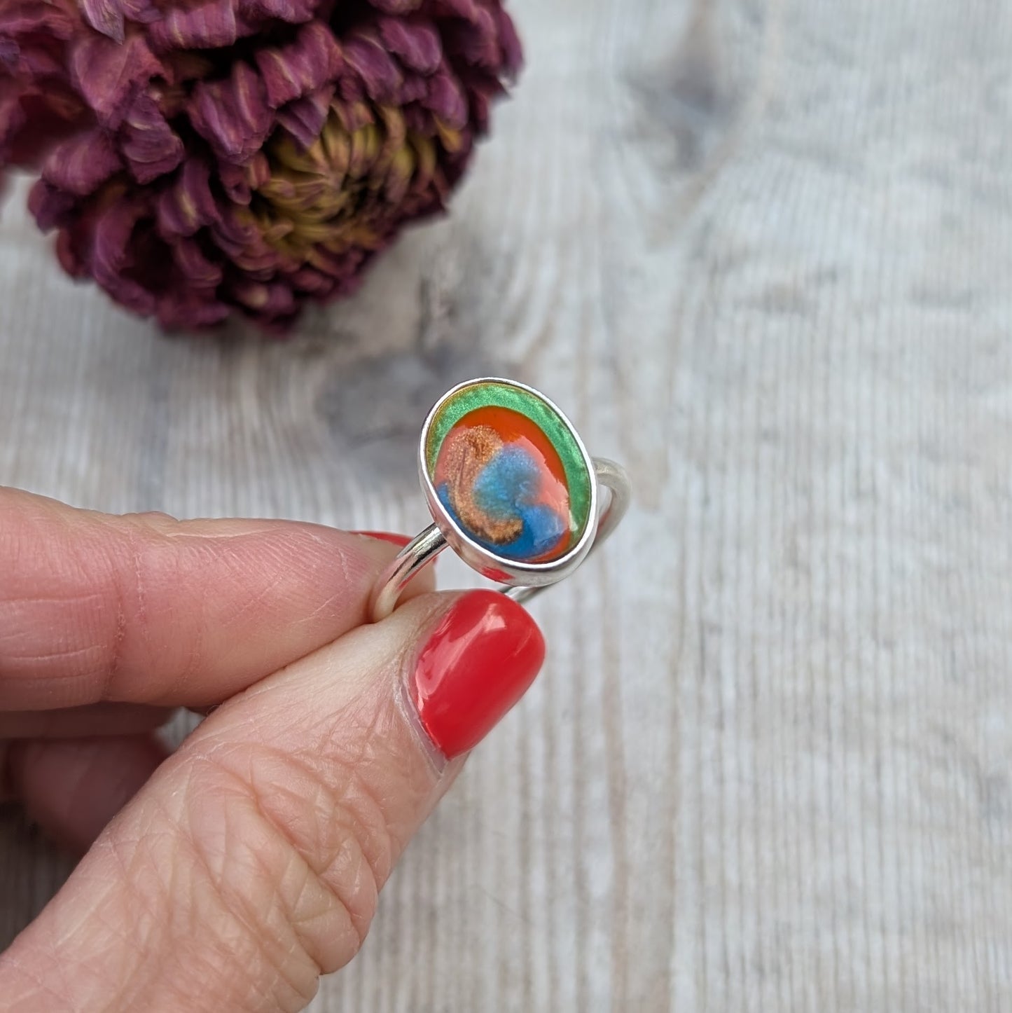 A close-up of the Surfite ring held between fingers, showing the green, orange, copper and blue colours in the stone.