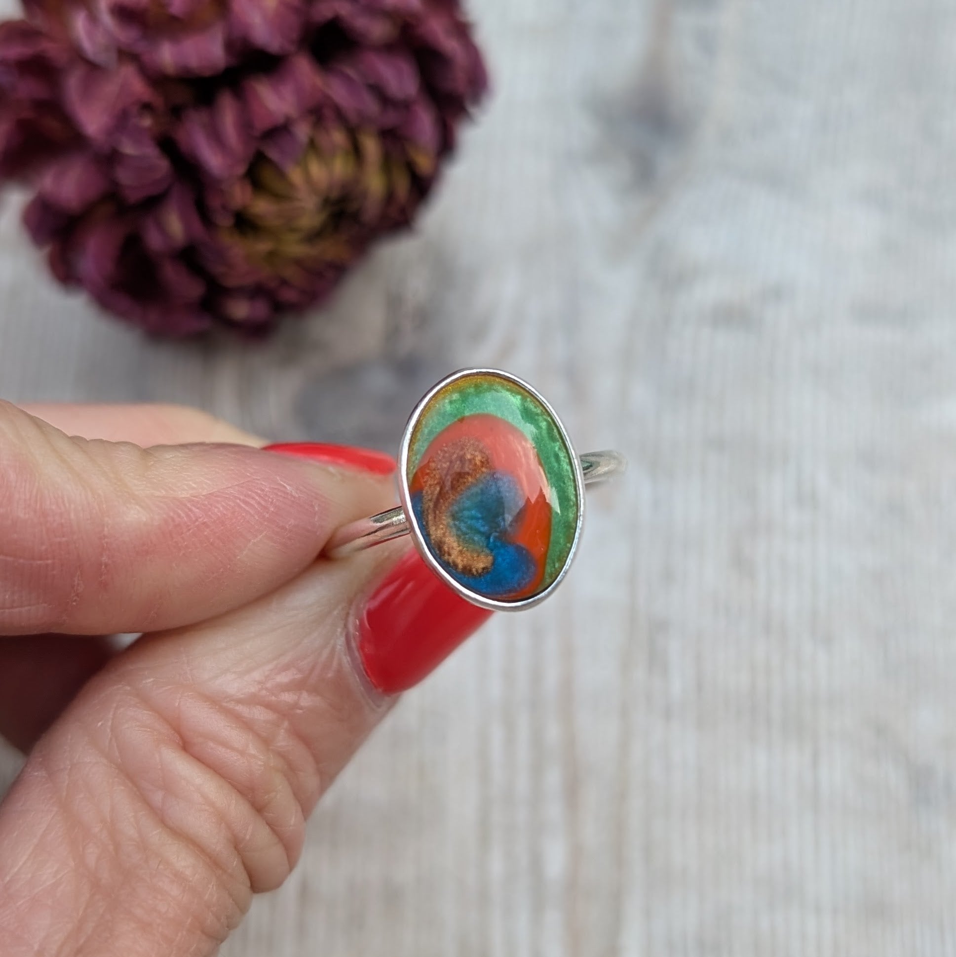 A close-up of the Surfite ring held between fingers, showing the green, orange, copper and blue colours in the stone.