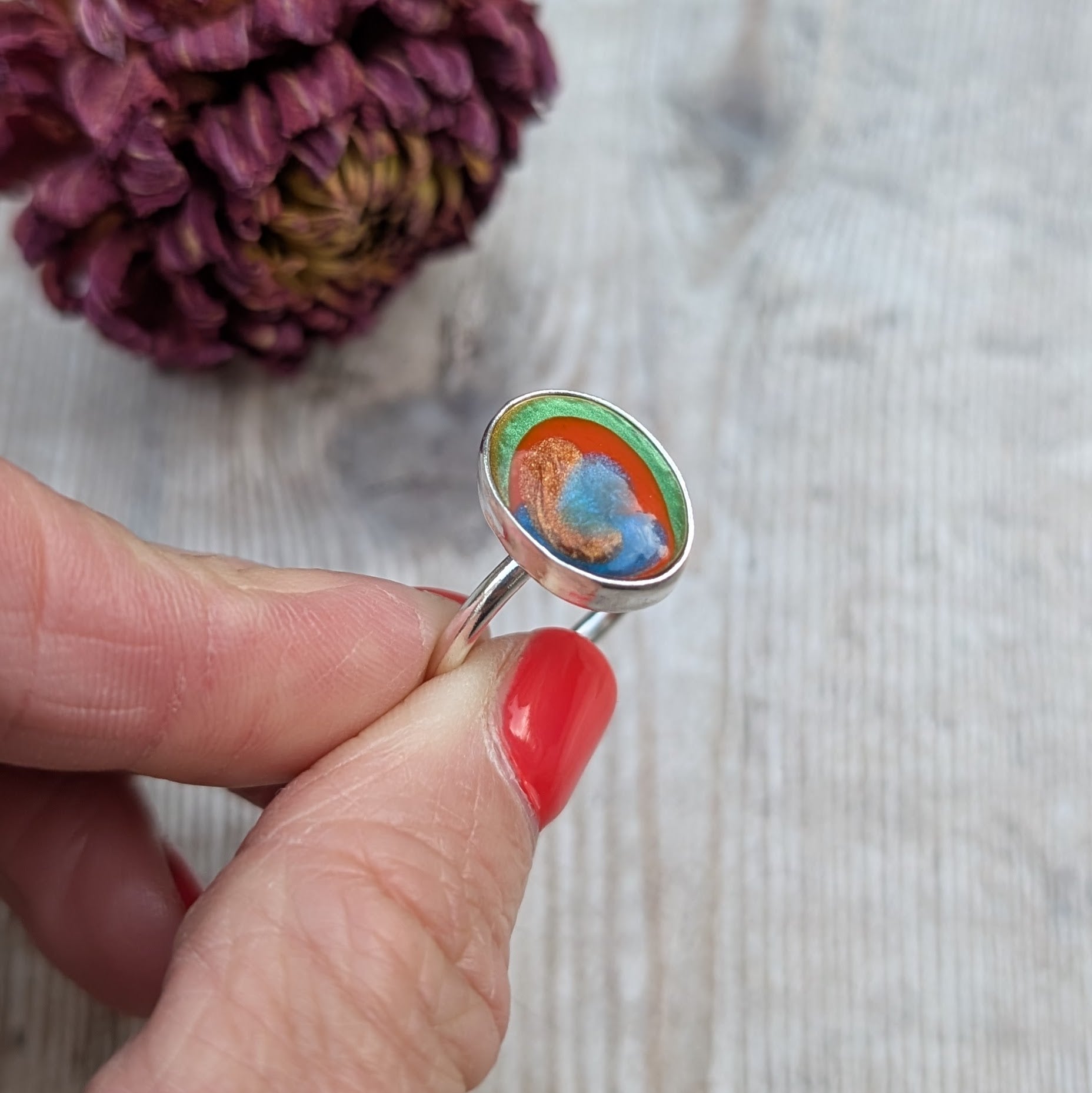 A close-up of the Surfite ring held between fingers, showing the green, orange, copper and blue colours in the stone.