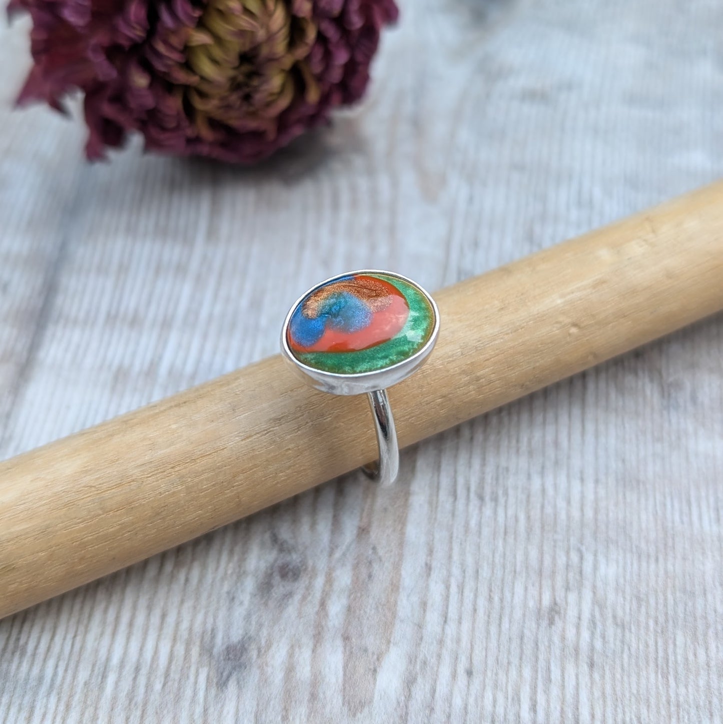 The ring displayed on a wooden ring mandrel, with a textured light wooden background and dried flower behind.