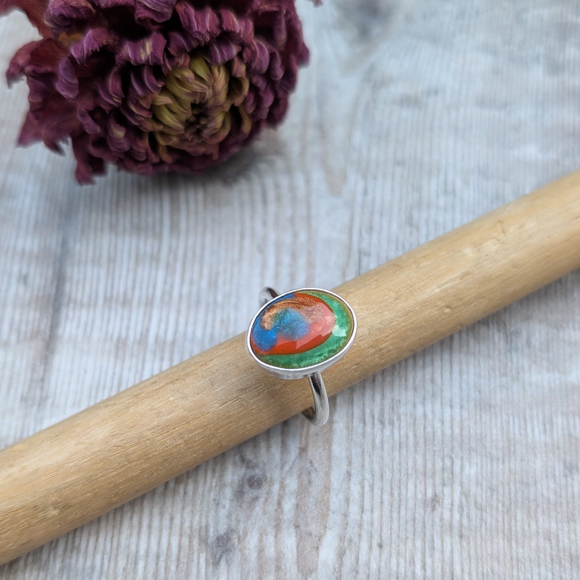 The ring displayed on a wooden ring mandrel, with a textured light wooden background and dried flower behind.