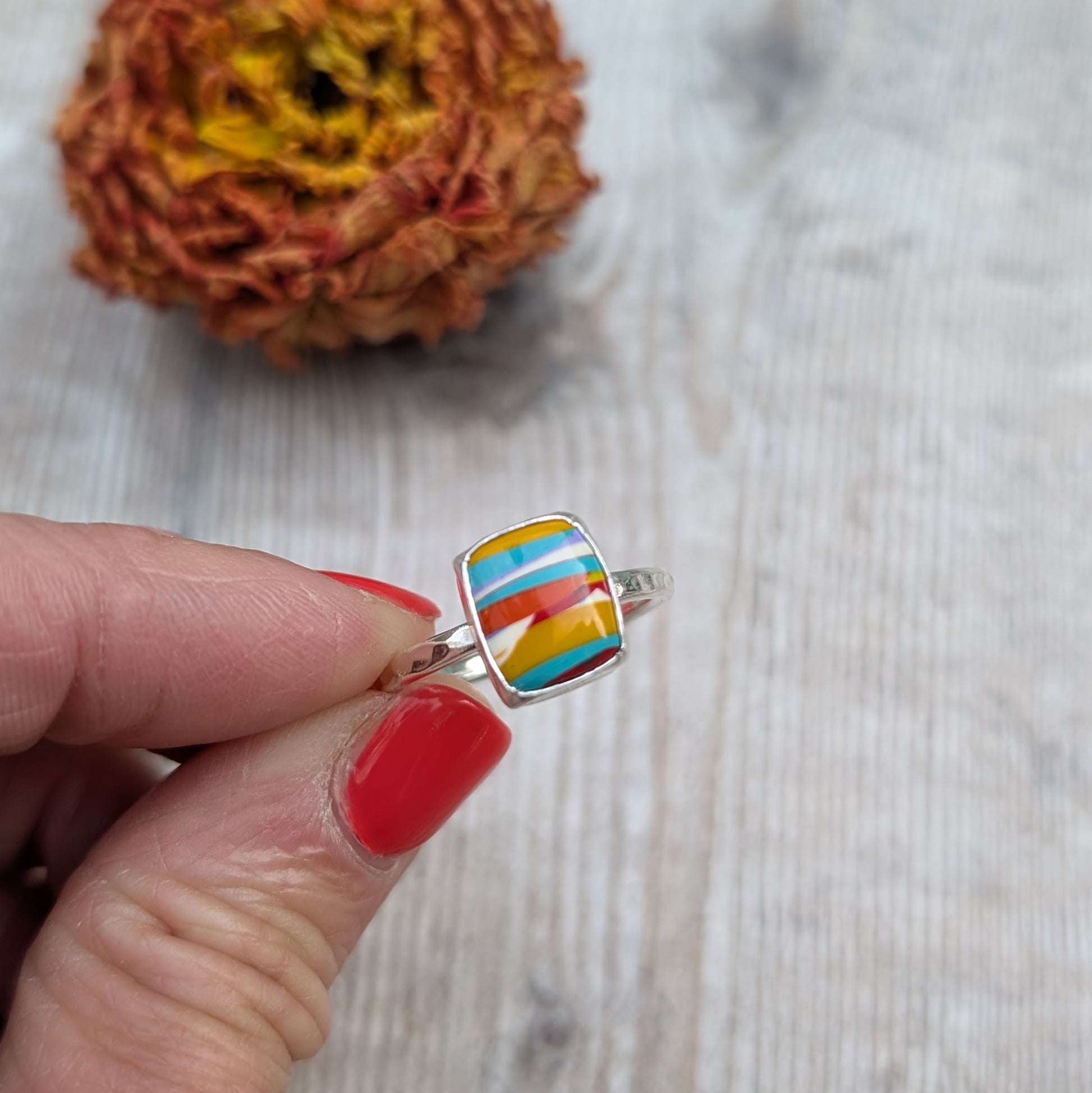 Close-up of a handmade silver ring with a colourful rectangular Surfite stone held between fingers with red nail polish.