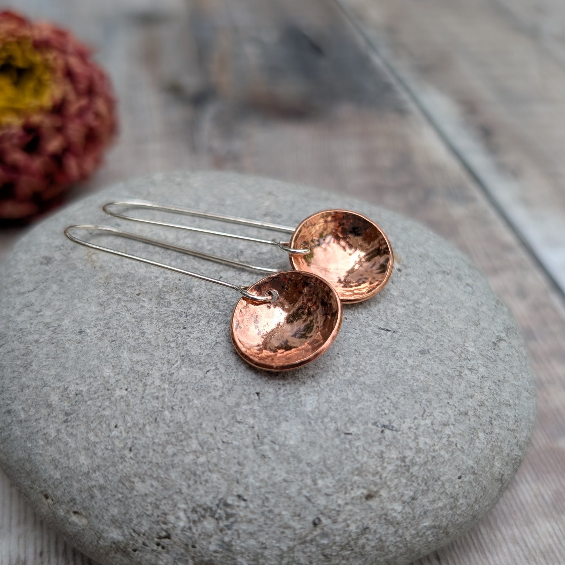 A pair of handmade hammered copper dome disc earrings with long silver hooks, displayed on a smooth grey stone with a blurred dried flower in the background.