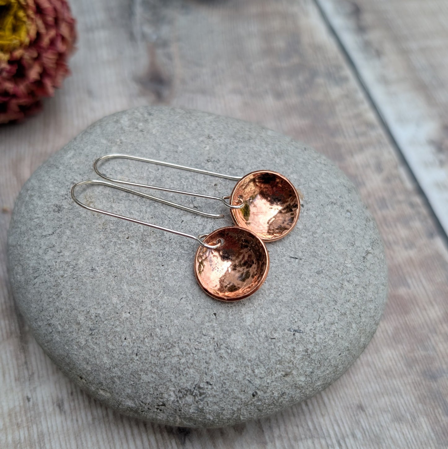 A pair of handmade hammered copper dome disc earrings with long silver hooks, displayed on a smooth grey stone with a blurred dried flower in the background.