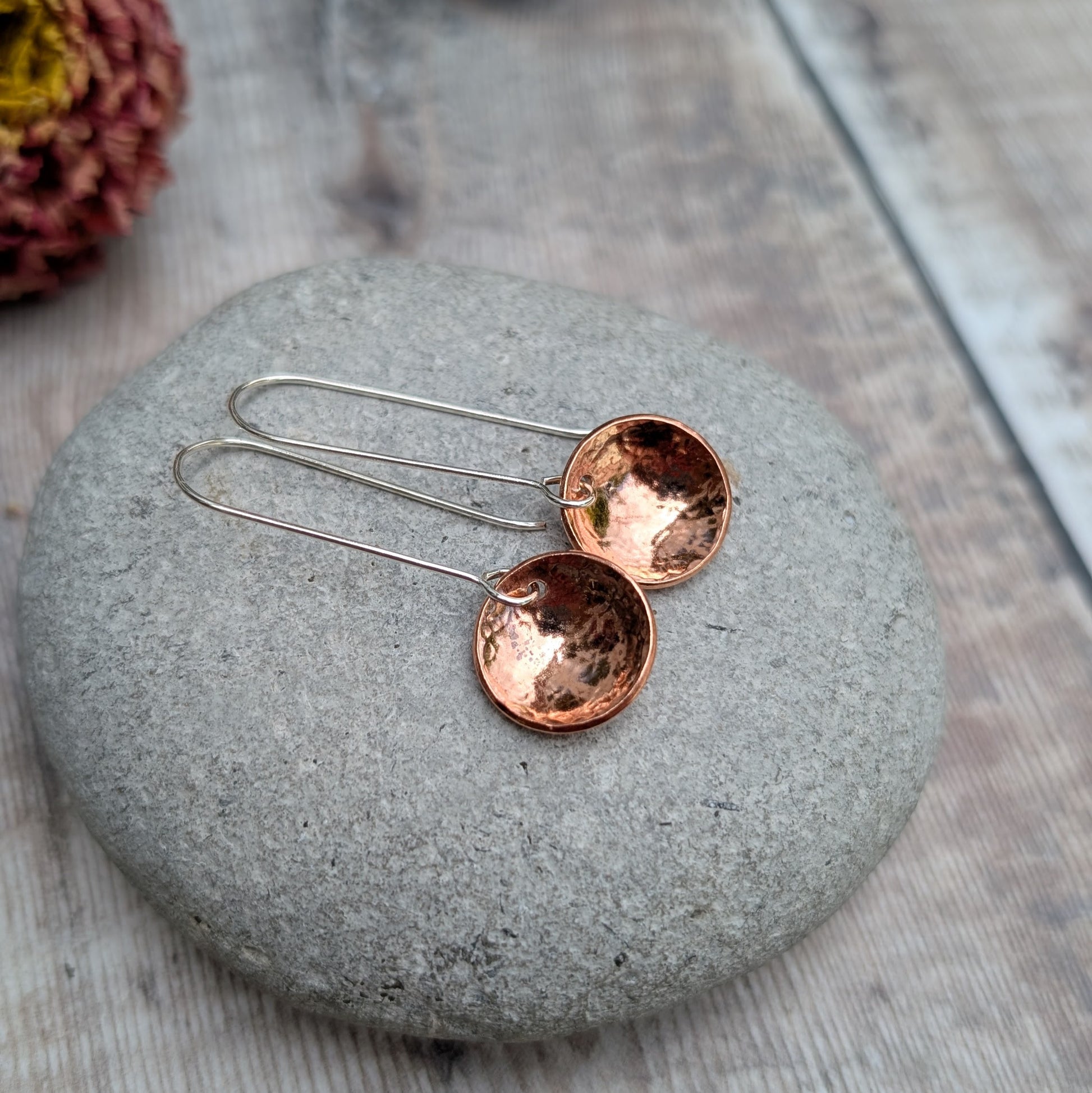 A pair of handmade hammered copper dome disc earrings with long silver hooks, displayed on a smooth grey stone with a blurred dried flower in the background.