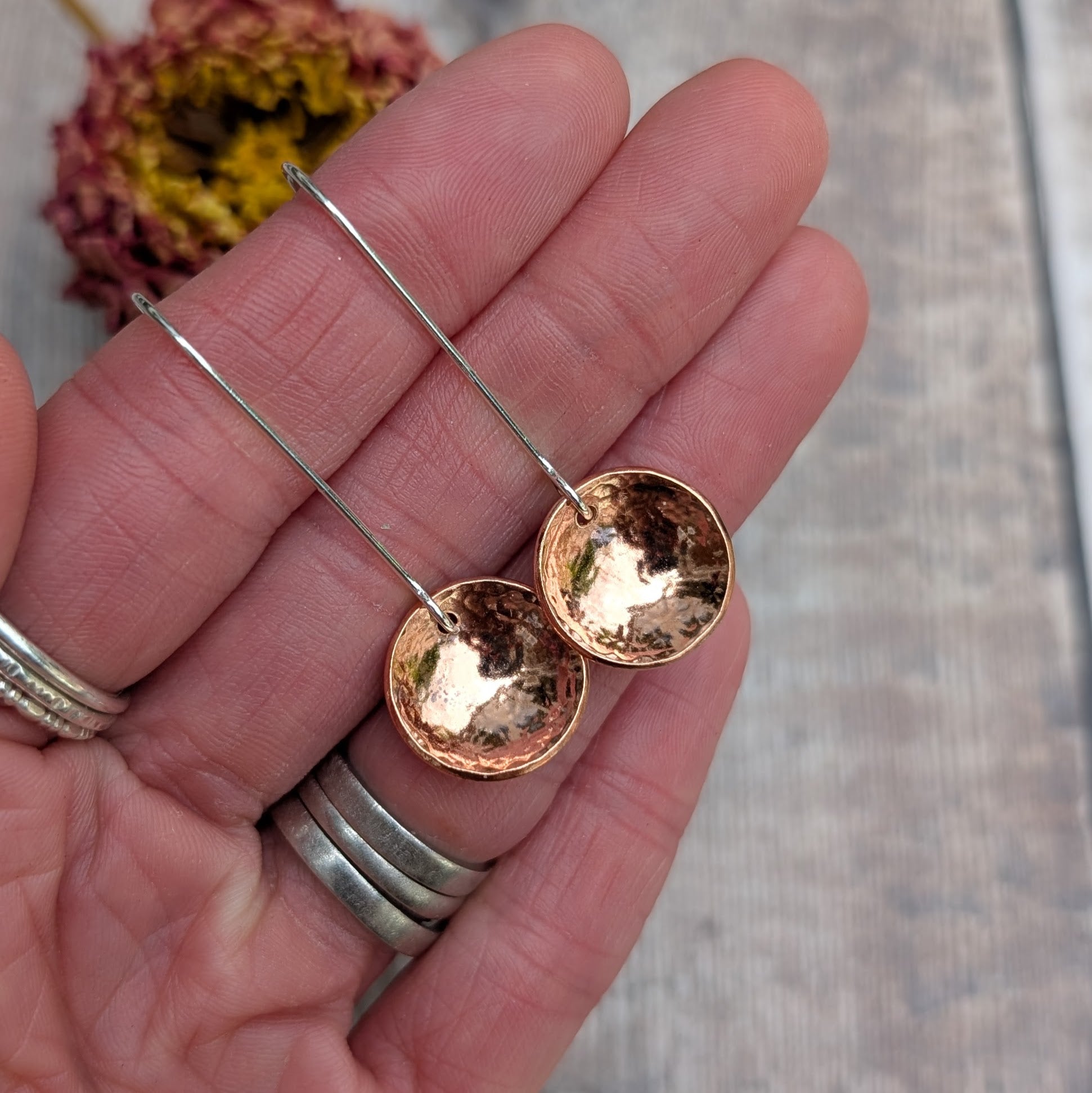 Close-up of a hand holding the hammered copper dome disc earrings by their long silver ear wires, showing the reflective, curved copper surface.
