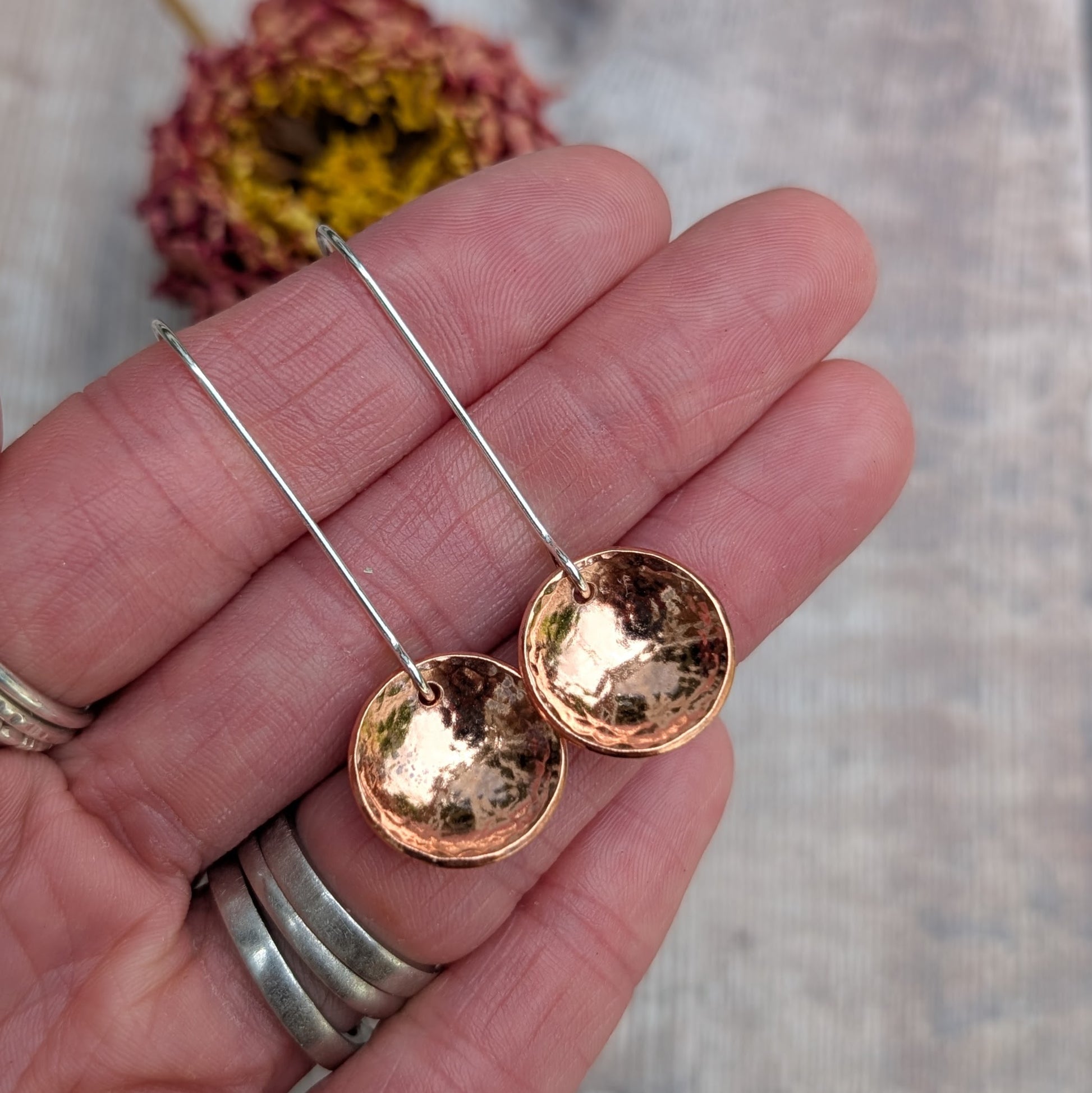 Close-up of a hand holding the hammered copper dome disc earrings by their long silver ear wires, showing the reflective, curved copper surface.