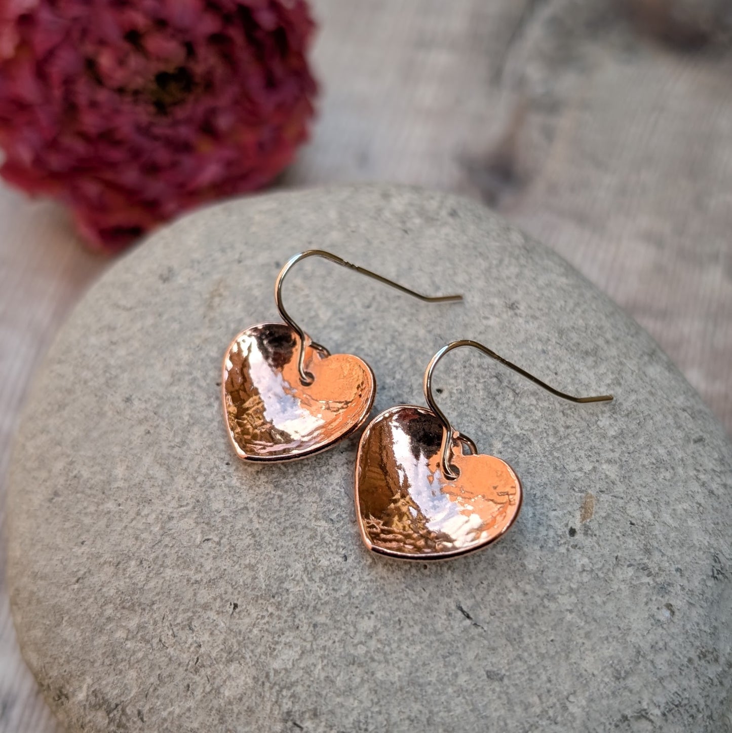 A pair of hammered copper heart earrings resting on a smooth grey stone, with a pink dried flower in the background.