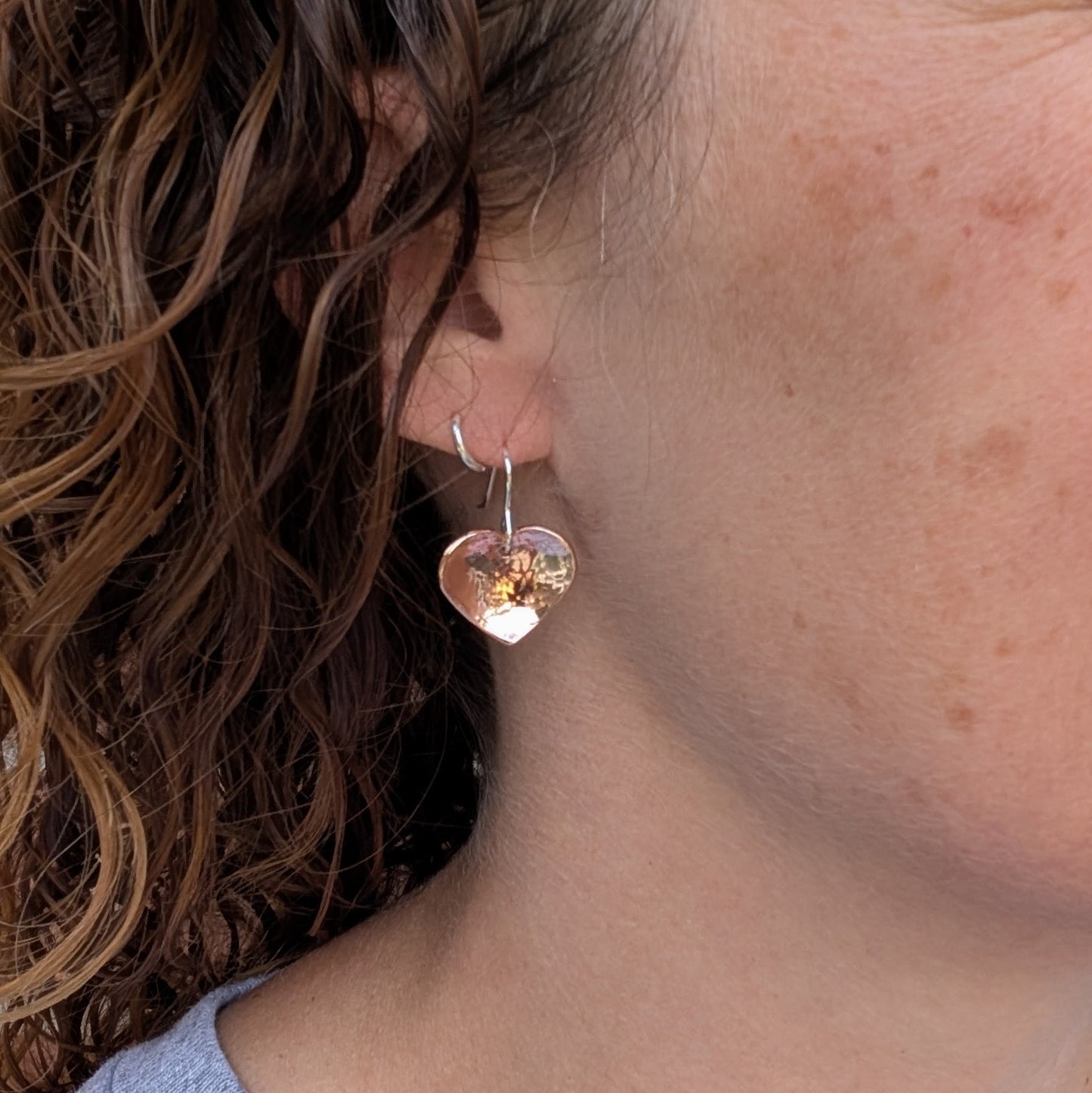 A hammered copper heart earring being worn, showing its size and drop style against the wearer’s ear.