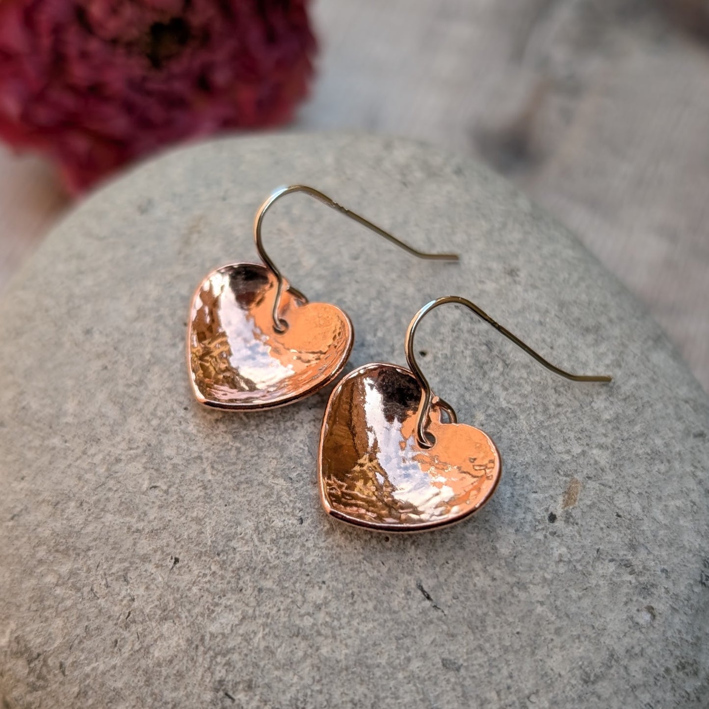 A pair of hammered copper heart earrings displayed on a grey stone surface with a soft-focus pink flower in the background.