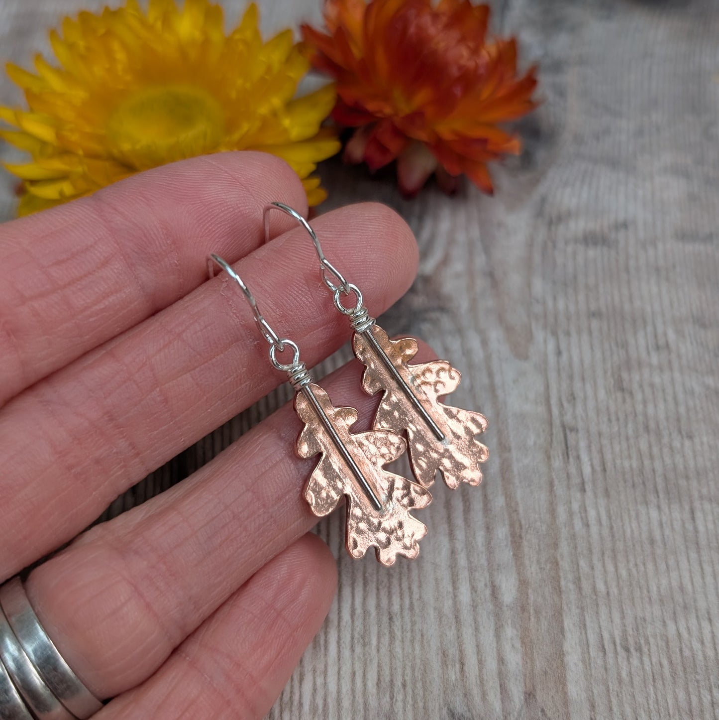 Copper hammered oak leaf earrings held between fingers, showing their shape, texture, and recycled sterling silver hooks.