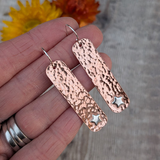 Copper hammered rectangle earrings with silver stars resting on a hand, showing scale, texture, and the contrast between copper and silver.