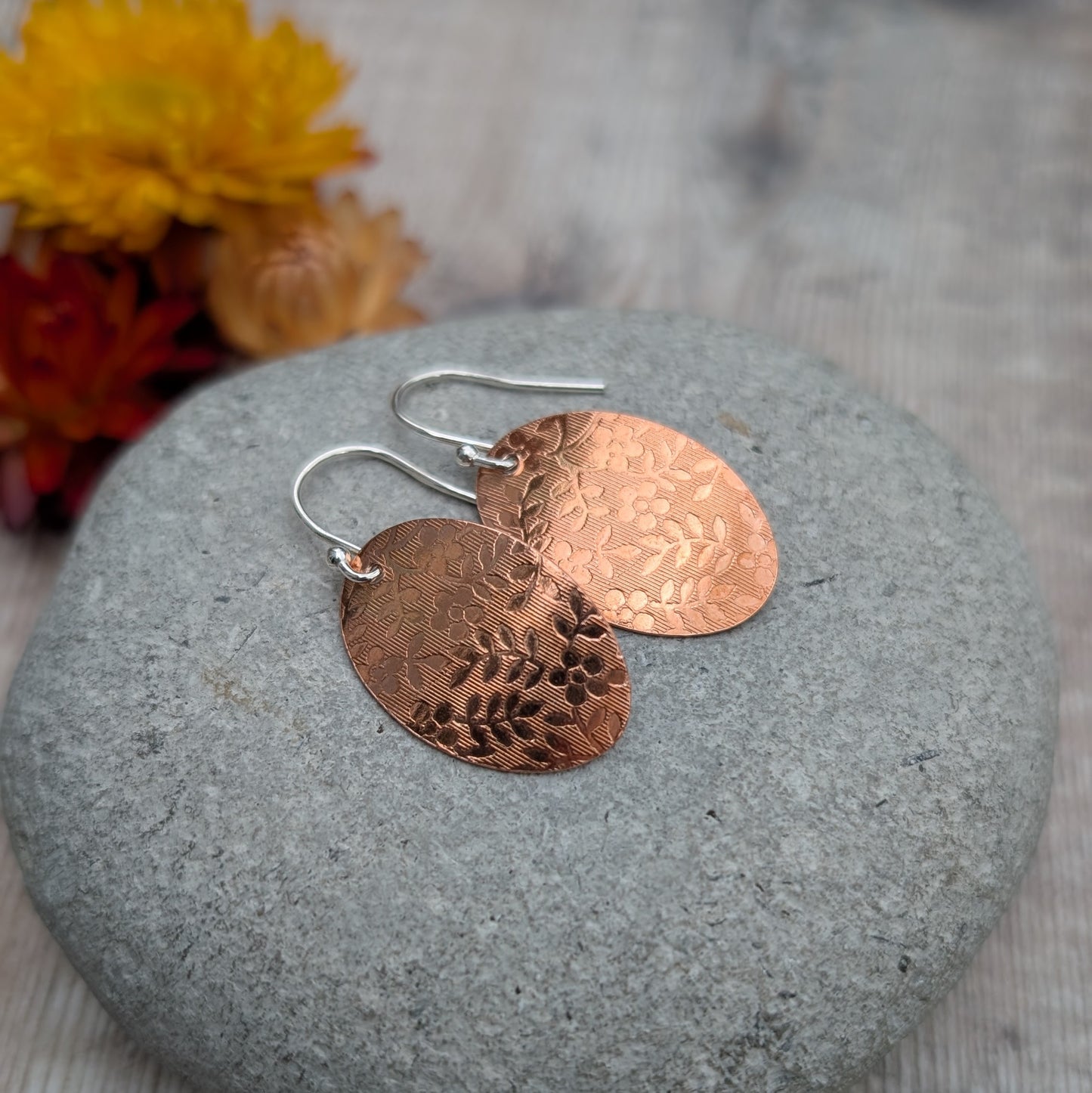 Copper floral pattern oval disc earrings displayed on a stone with dried flowers in the background.