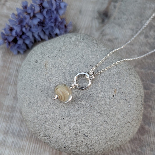 Cream lampwork and silver circle necklace displayed on a smooth grey stone with a soft purple floral background.