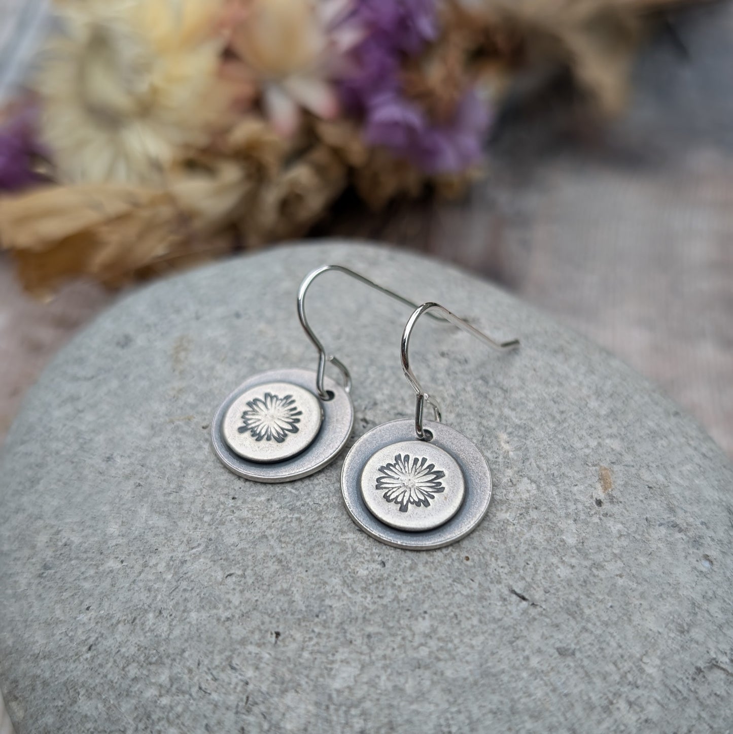 Close-up of oxidised silver earrings with a daisy pattern on each disc, displayed on a smooth stone with dried flowers in the background.