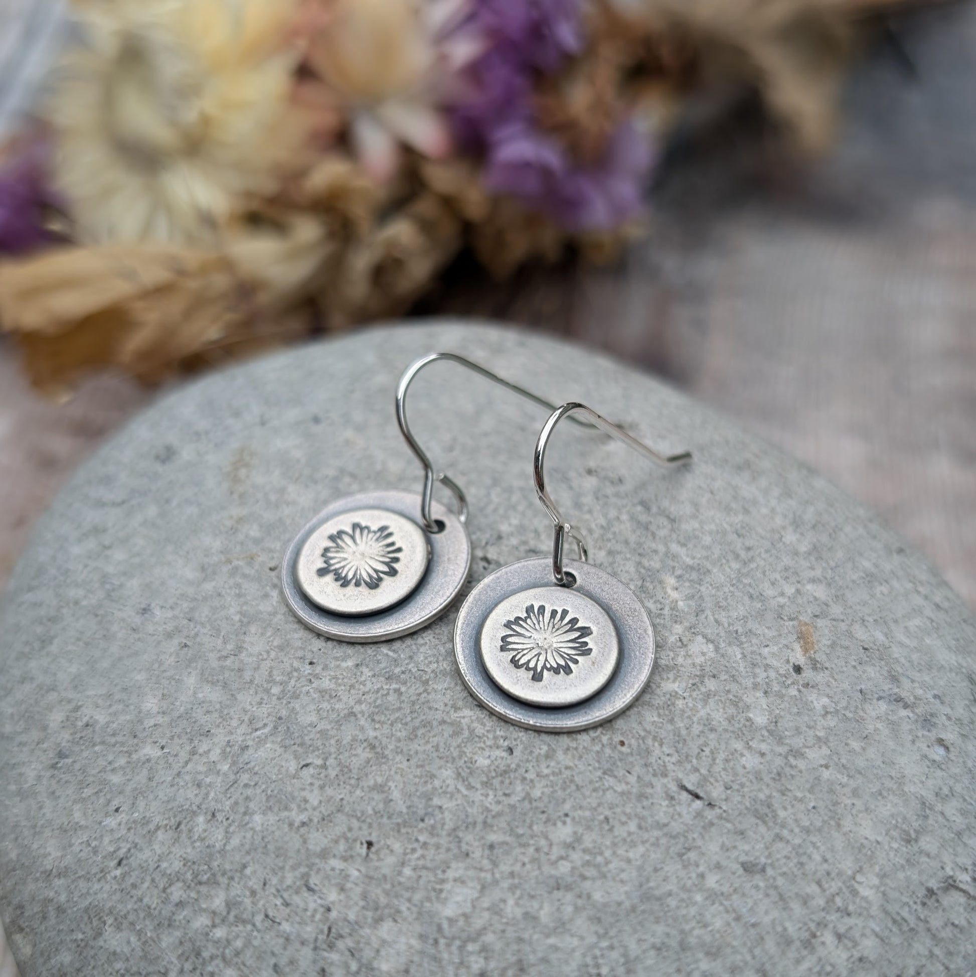 Close-up of oxidised silver earrings with a daisy pattern on each disc, displayed on a smooth stone with dried flowers in the background.