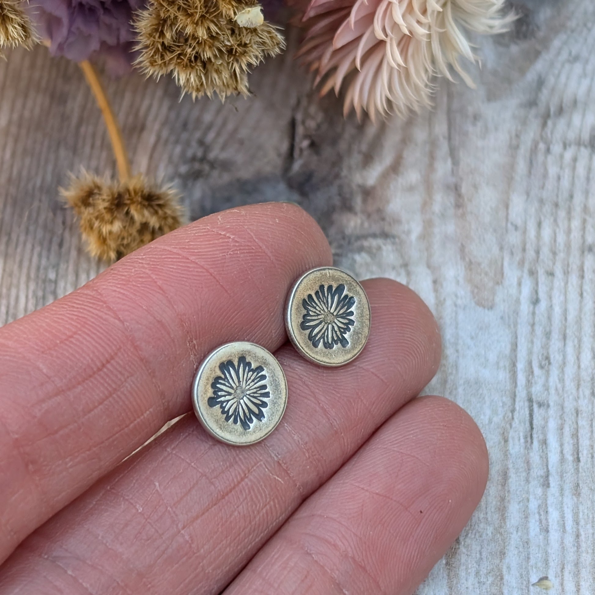 A pair of silver stud earrings with an oxidised daisy pattern on circular discs, held between fingers. Dried flowers in soft purple and beige tones are blurred in the background.