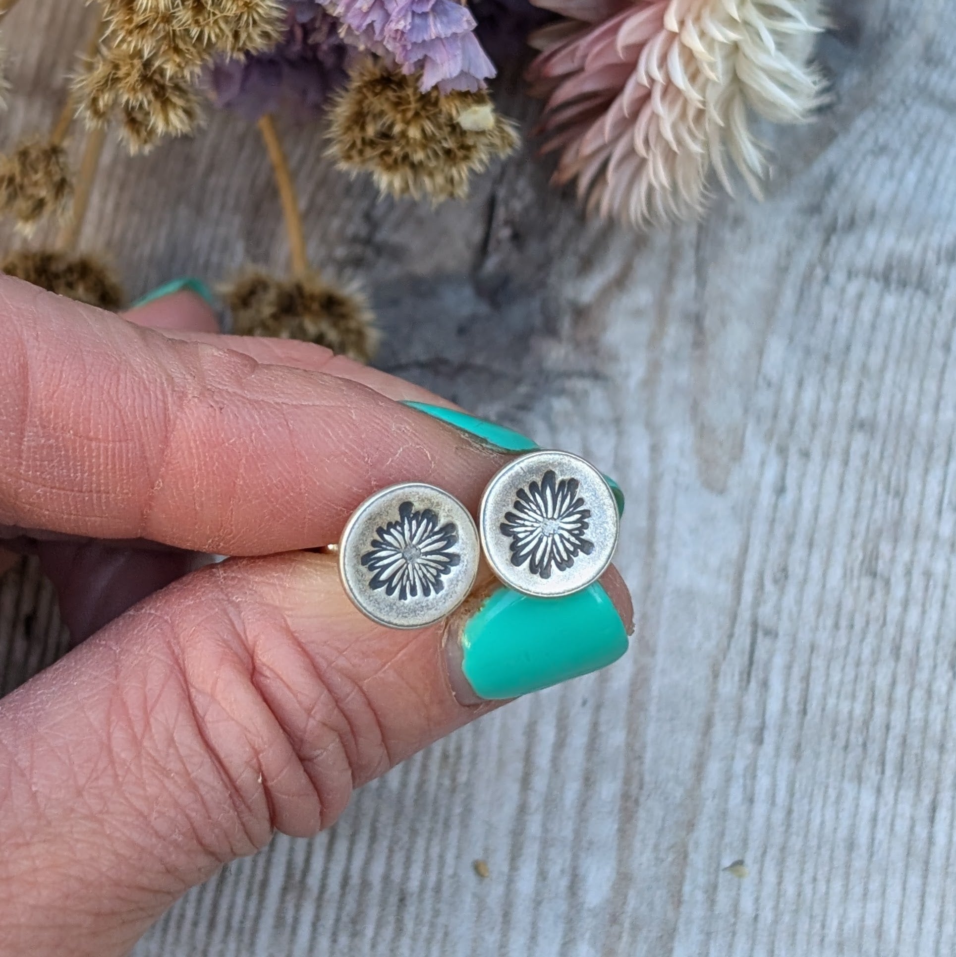 A pair of silver stud earrings with an oxidised daisy pattern on circular discs, held between fingers. Dried flowers in soft purple and beige tones are blurred in the background.
