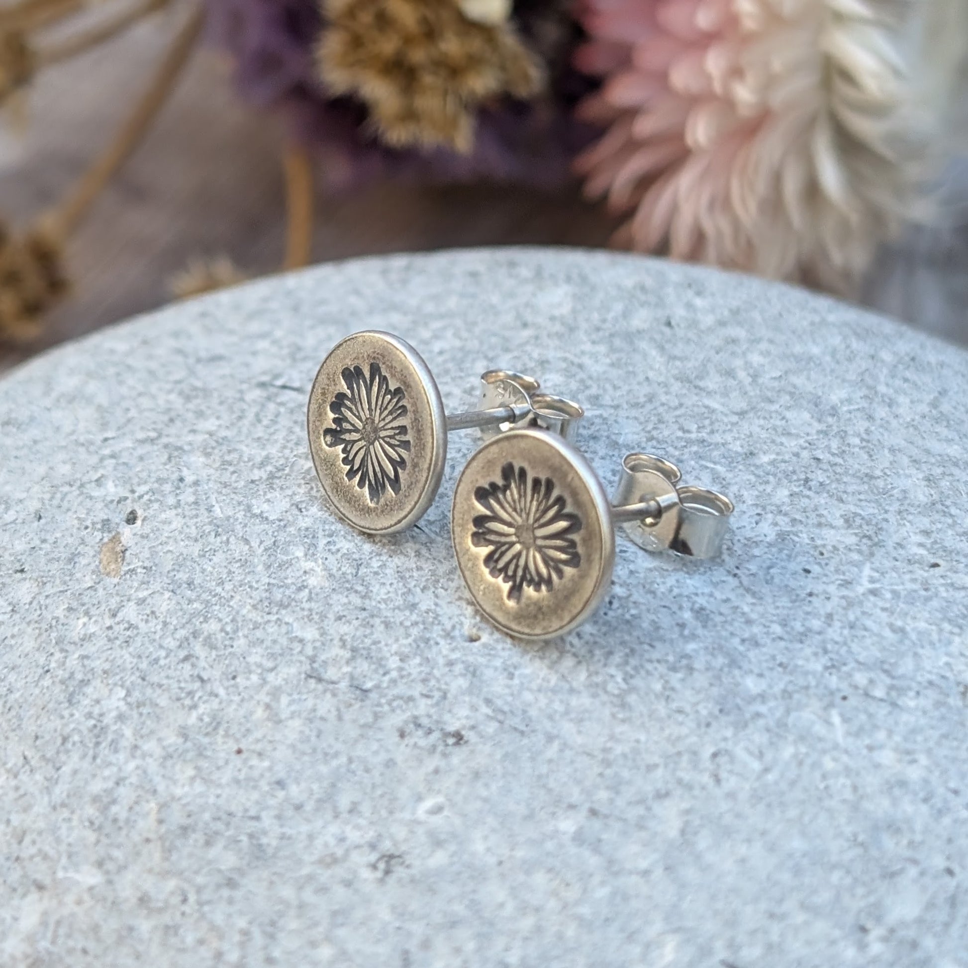 A close-up of silver stud earrings with an oxidised daisy pattern impressed on circular discs, resting on a textured stone surface with blurred dried flowers in the background.