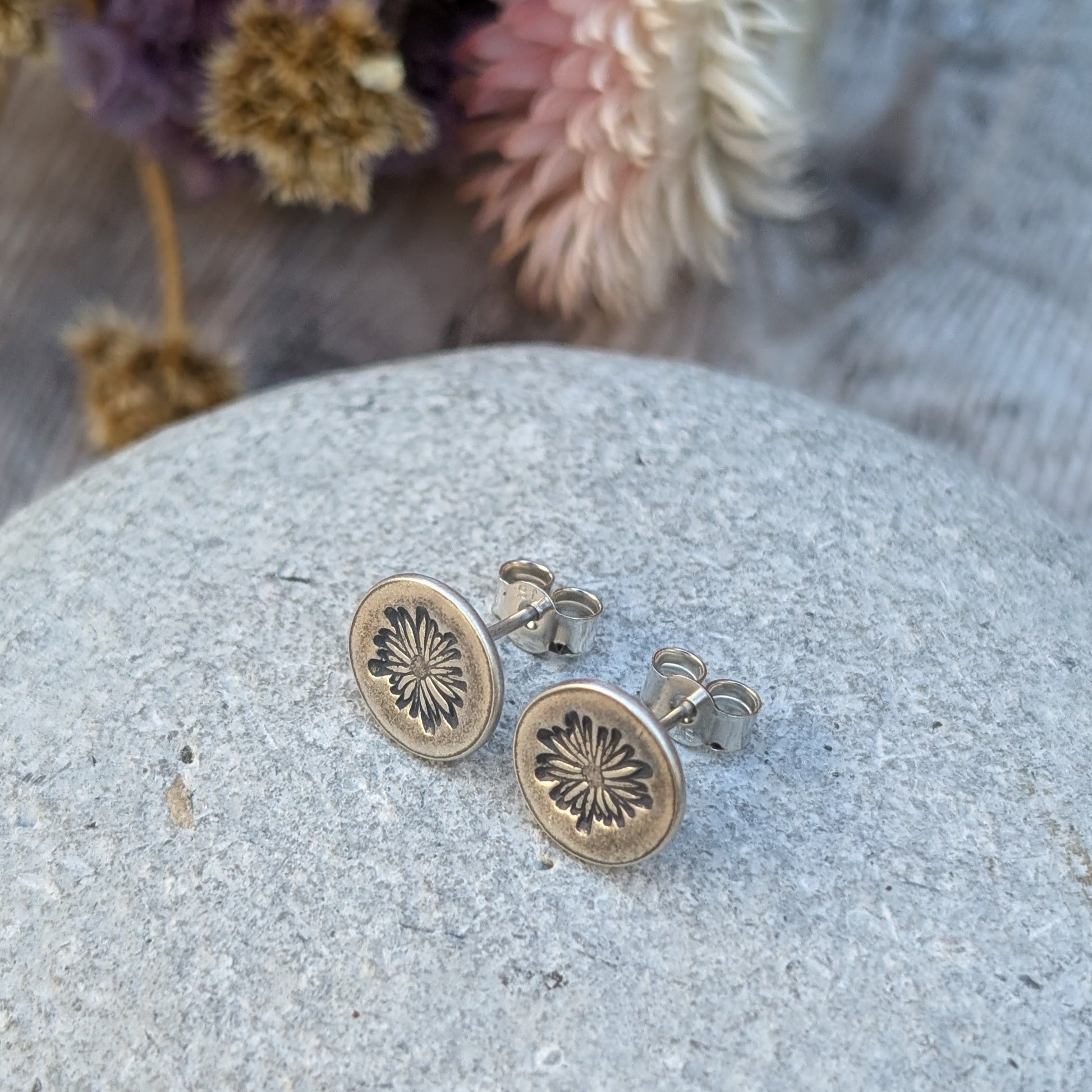 A close-up of silver stud earrings with an oxidised daisy pattern impressed on circular discs, resting on a textured stone surface with blurred dried flowers in the background.