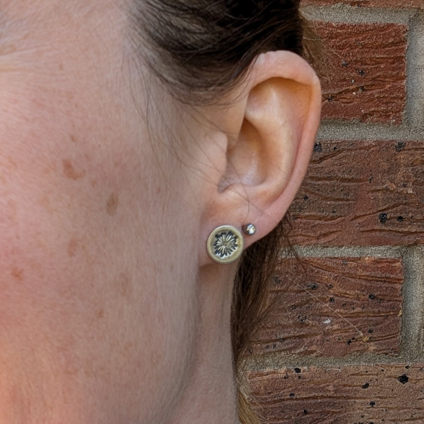 A silver daisy disc stud earring worn on the earlobe, with the detailed flower pattern standing out against the oxidised silver finish. A second small stud eA silver daisy disc stud earring worn on the earlobe, with the detailed flower pattern standing out against the oxidised silver finish. A second small stud earring is visible in the upper ear piercing, with a brick wall in the background.arring is visible in the upper ear piercing, with a brick wall in the background.
