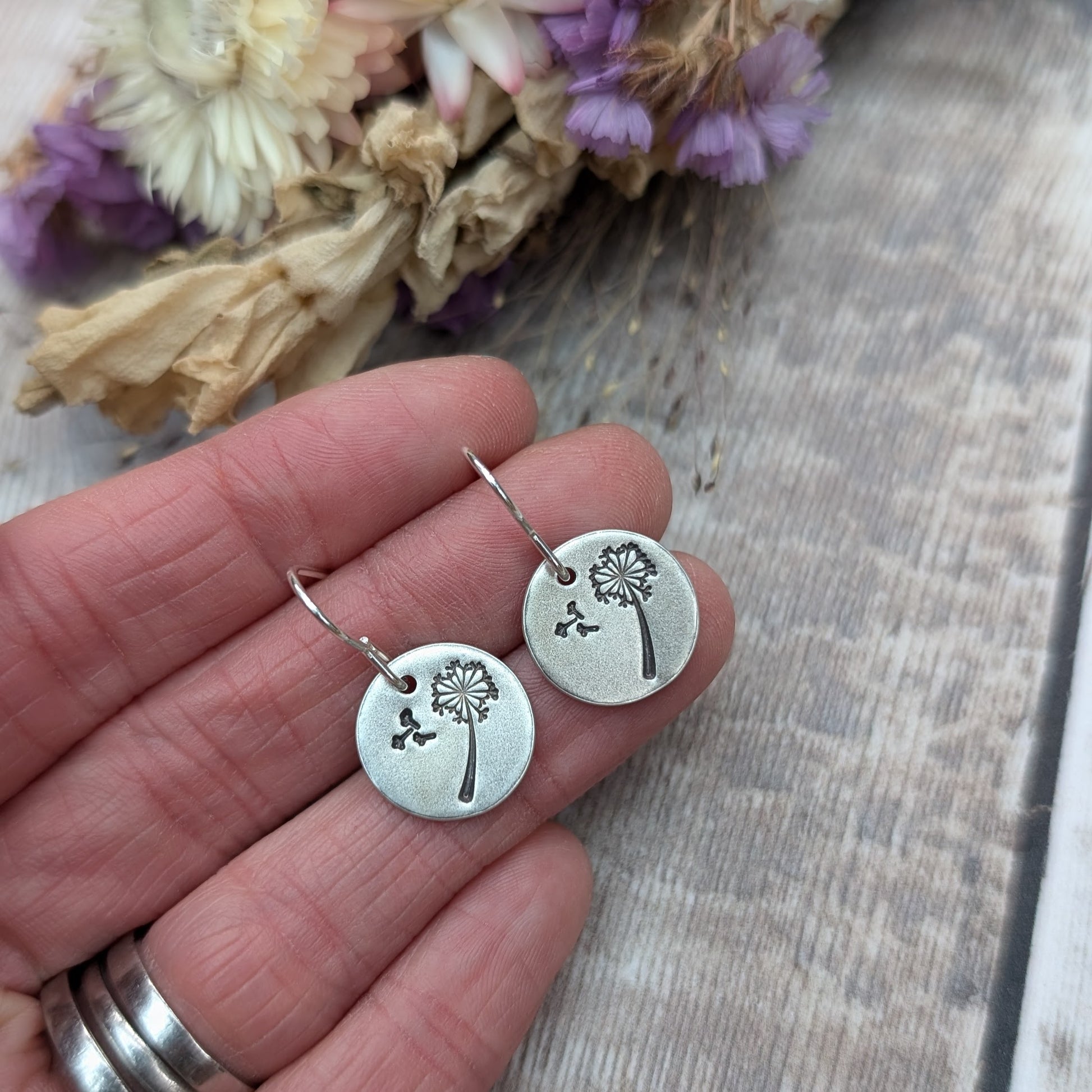 Close-up of a hand holding a pair of silver dangle earrings with a dandelion design etched onto circular discs, with dried flowers in the background.
