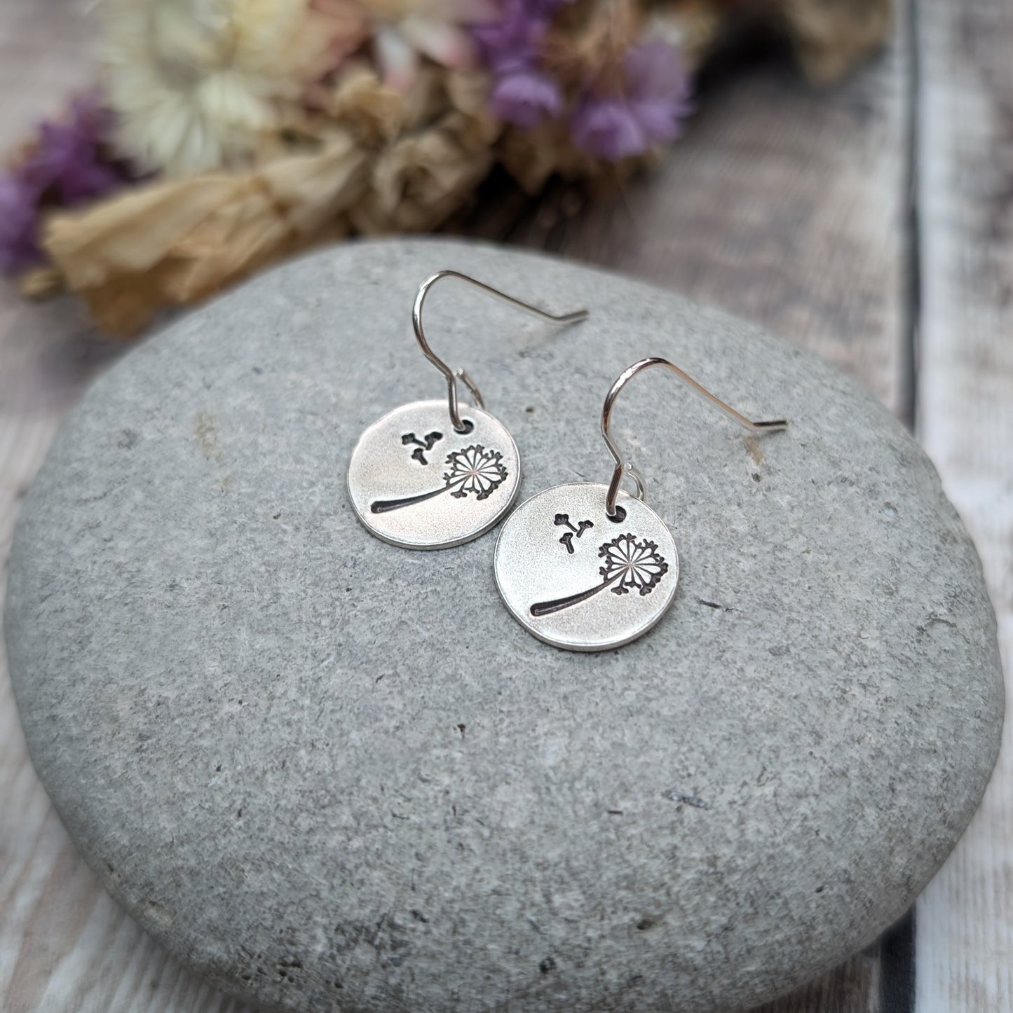 A pair of silver dangle earrings with a dandelion design etched onto circular discs, resting on a smooth stone with dried flowers in the background.