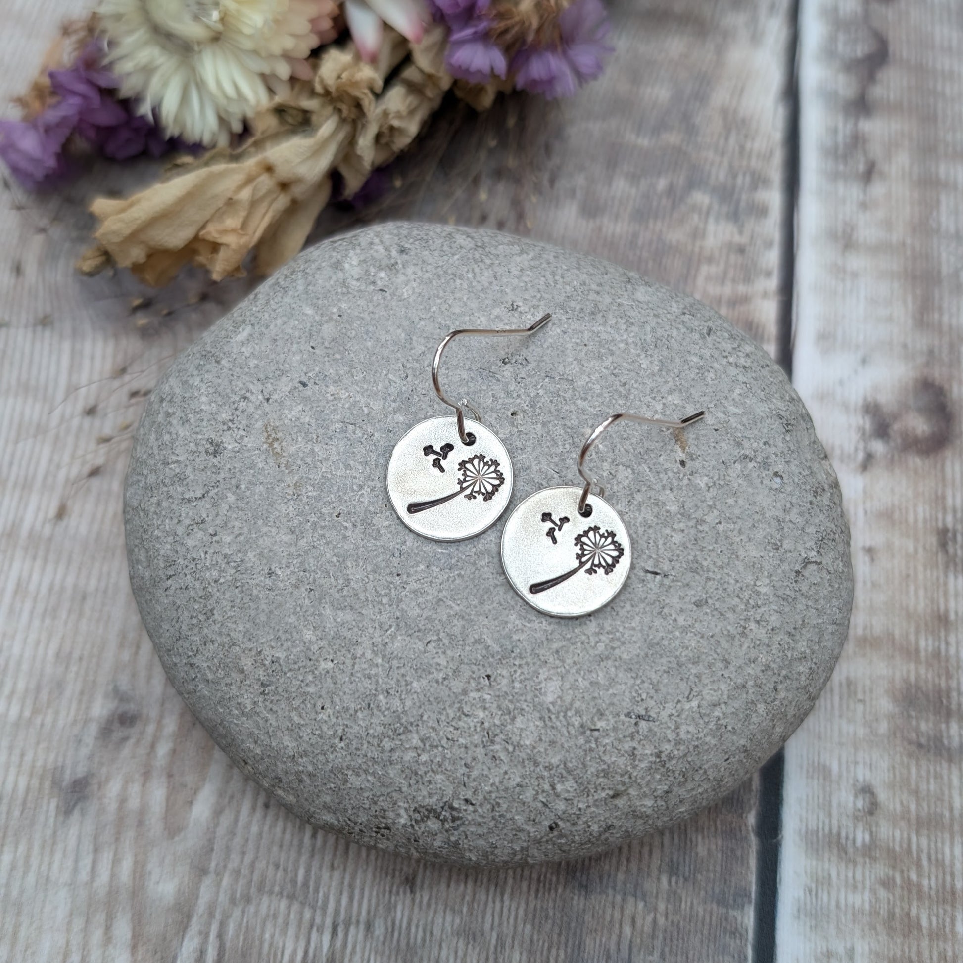 A pair of silver dangle earrings with a dandelion design etched onto circular discs, resting on a smooth stone with dried flowers in the background.