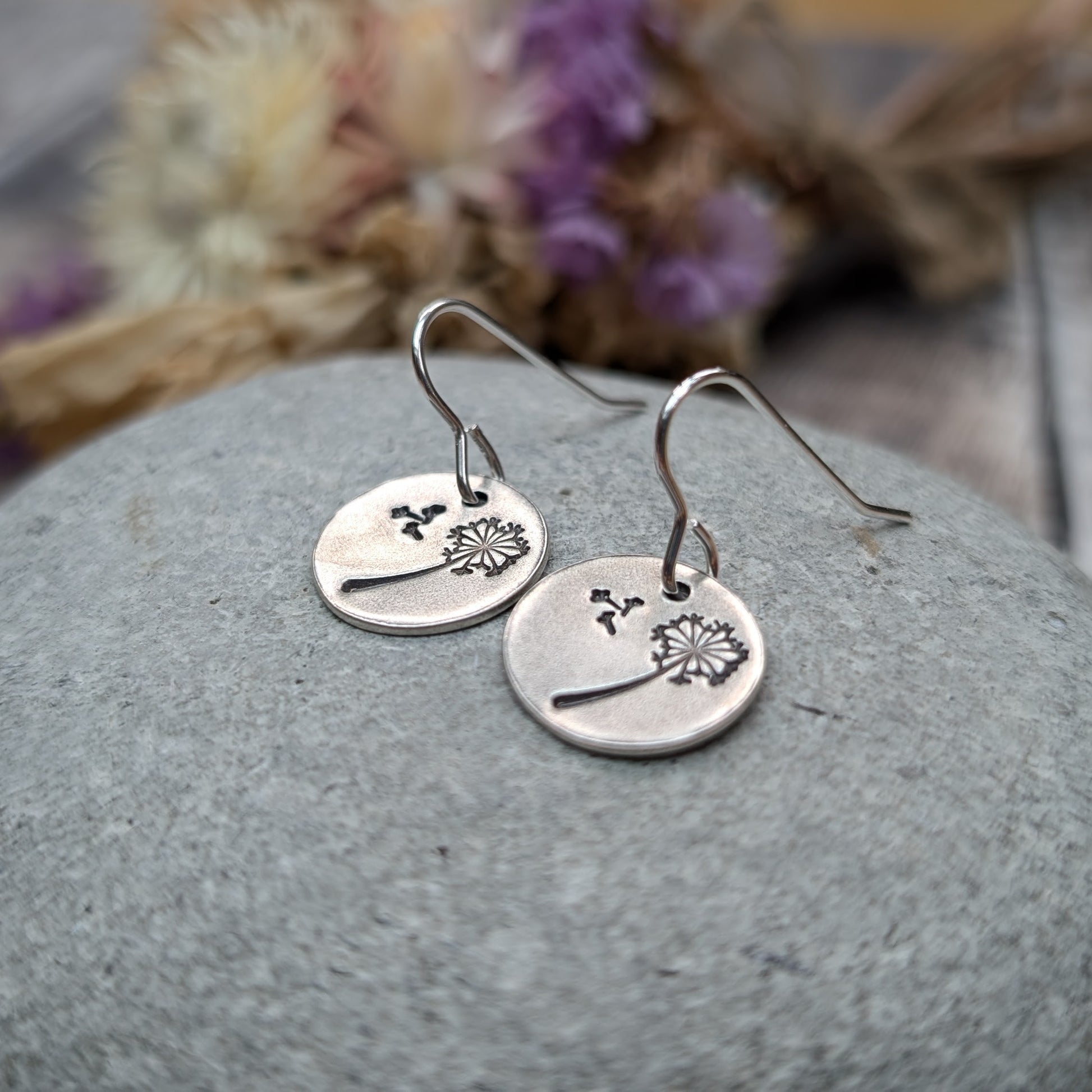 A pair of silver dangle earrings with a dandelion design etched onto circular discs, resting on a smooth stone with dried flowers in the background.