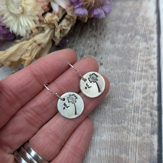 Close-up of a hand holding a pair of silver dangle earrings with a dandelion design etched onto circular discs, with dried flowers in the background.