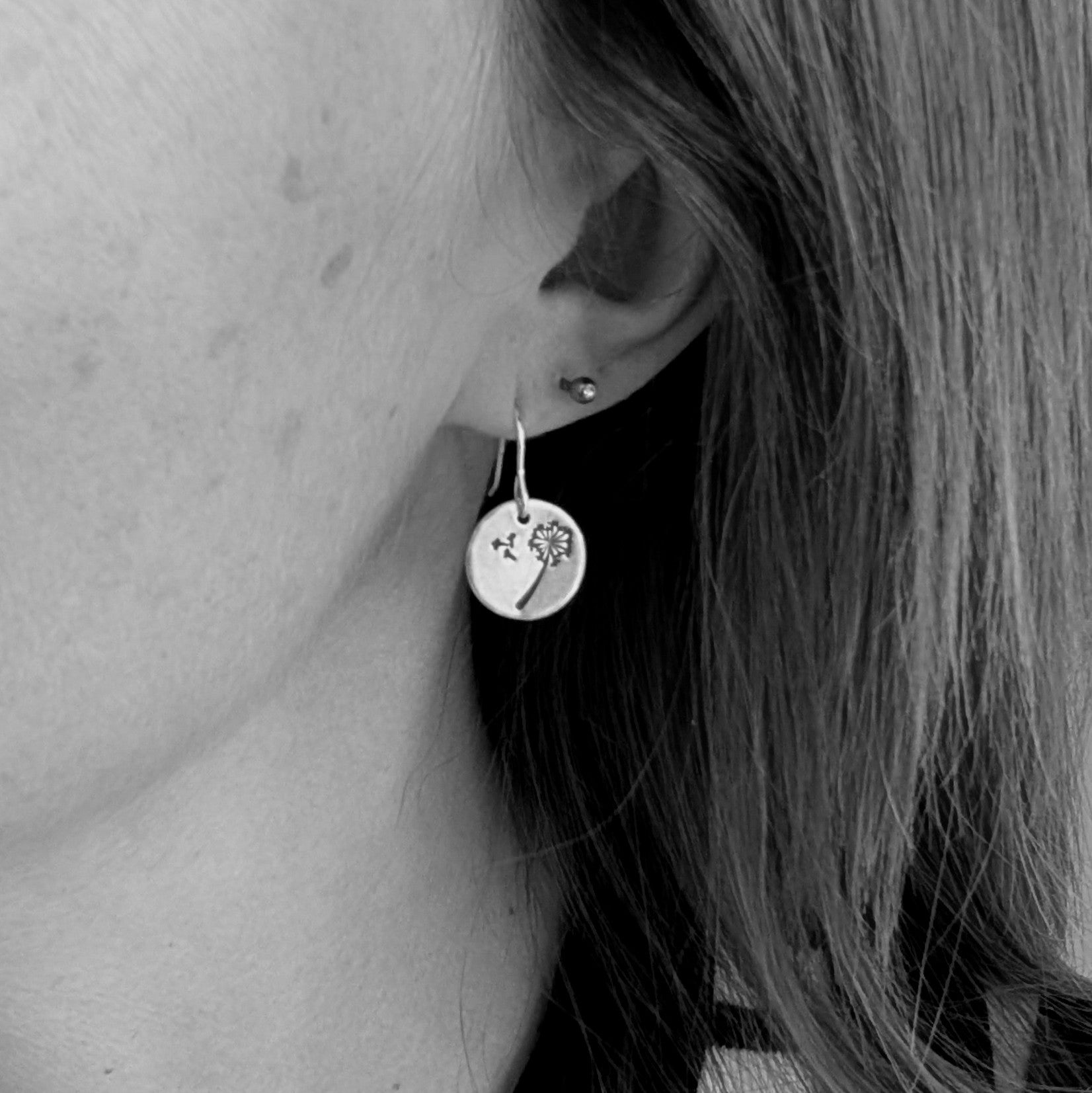 Black and white close-up of a woman wearing a single silver dangle earring with a dandelion design etched onto a circular disc.