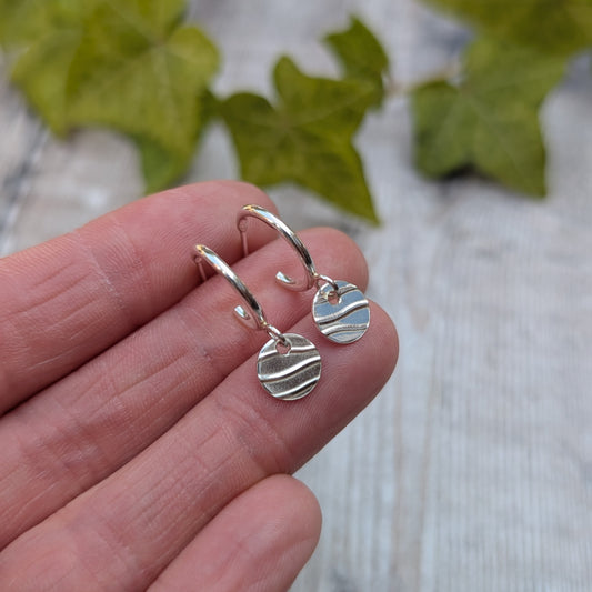 Close up of silver disc drop hoop earrings handmade from recycled Sterling Silver, shown resting on fingertips to highlight the hand stamped wave pattern.