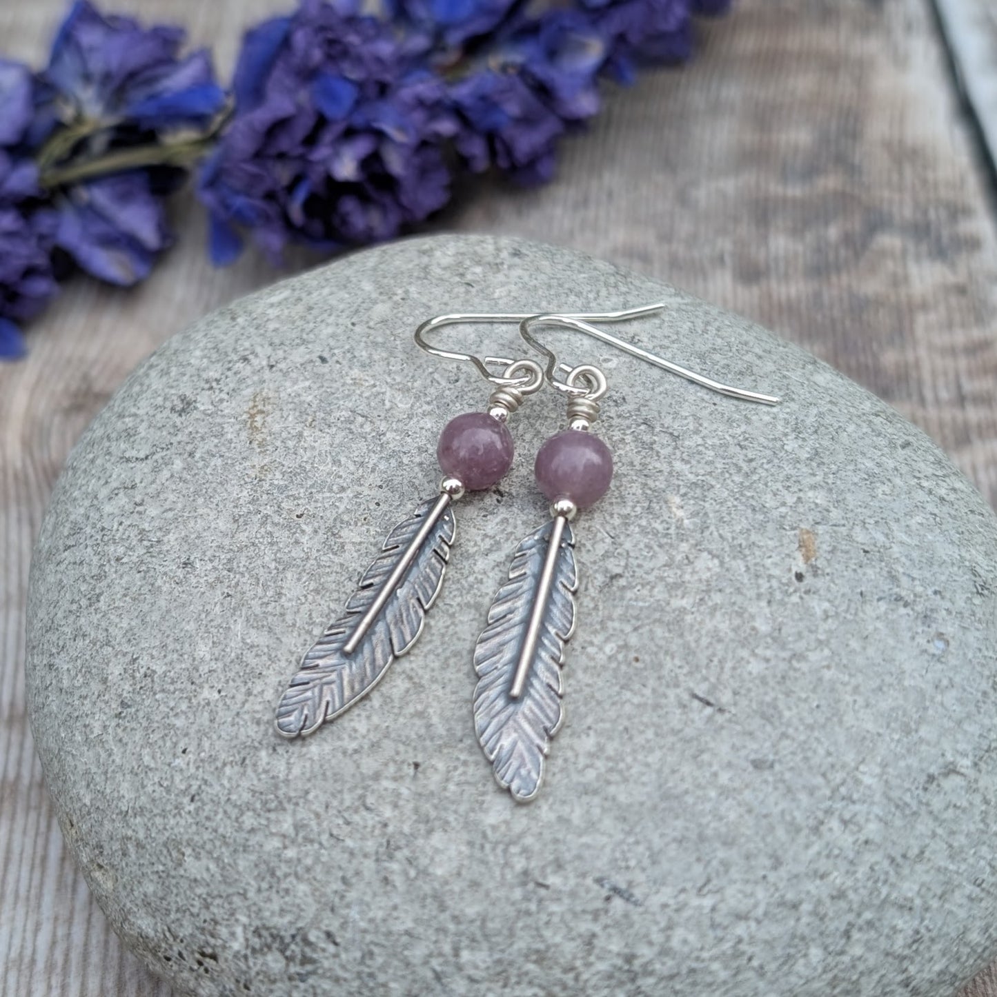 A pair of sterling silver feather earrings with lavender amethyst beads resting on a smooth stone, with dried purple flowers in the background, enhancing the natural aesthetic of the jewellery.