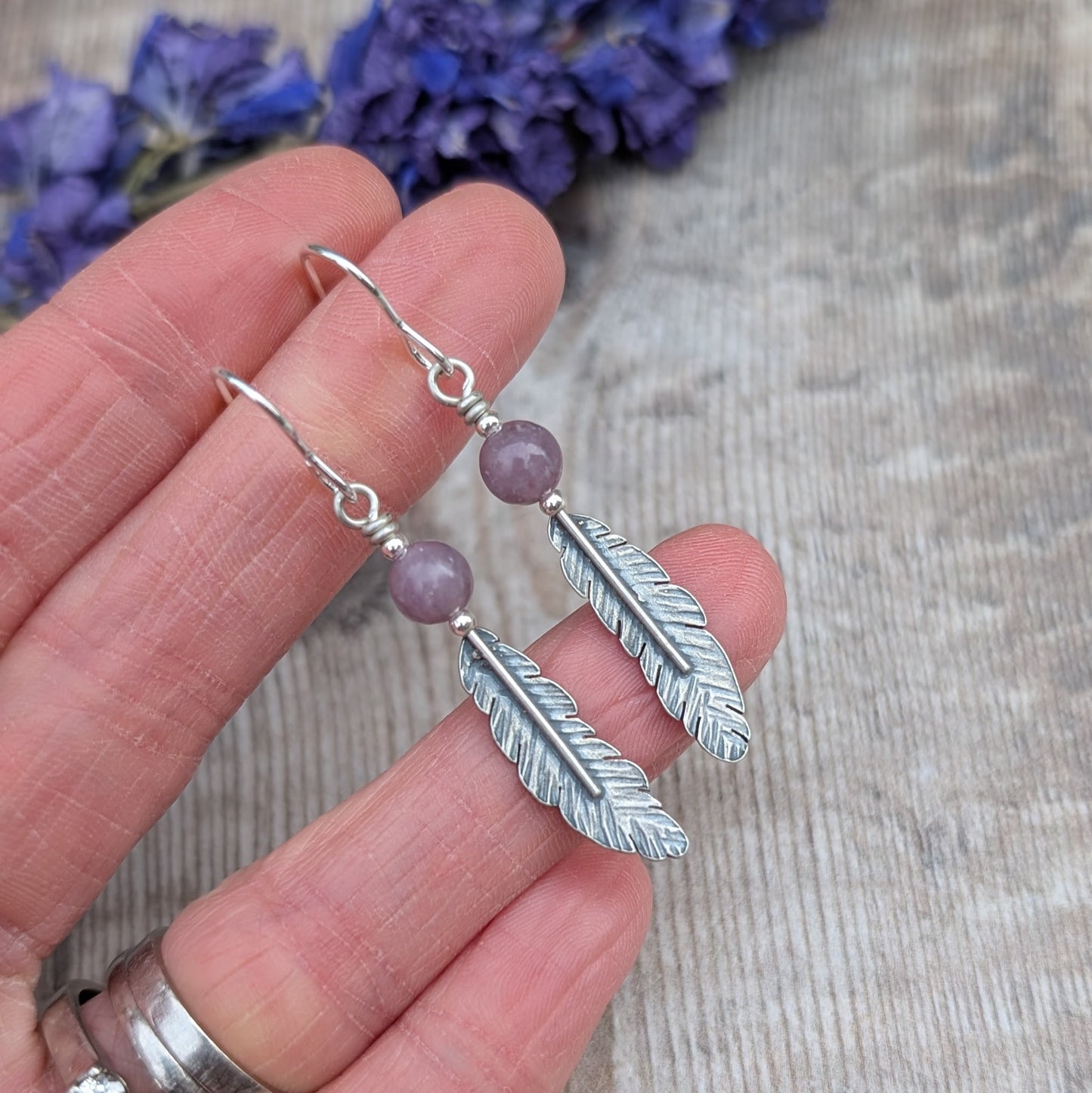 A close-up of a hand holding a pair of sterling silver feather earrings with lavender amethyst beads, showing the intricate feather texture and oxidised finish.