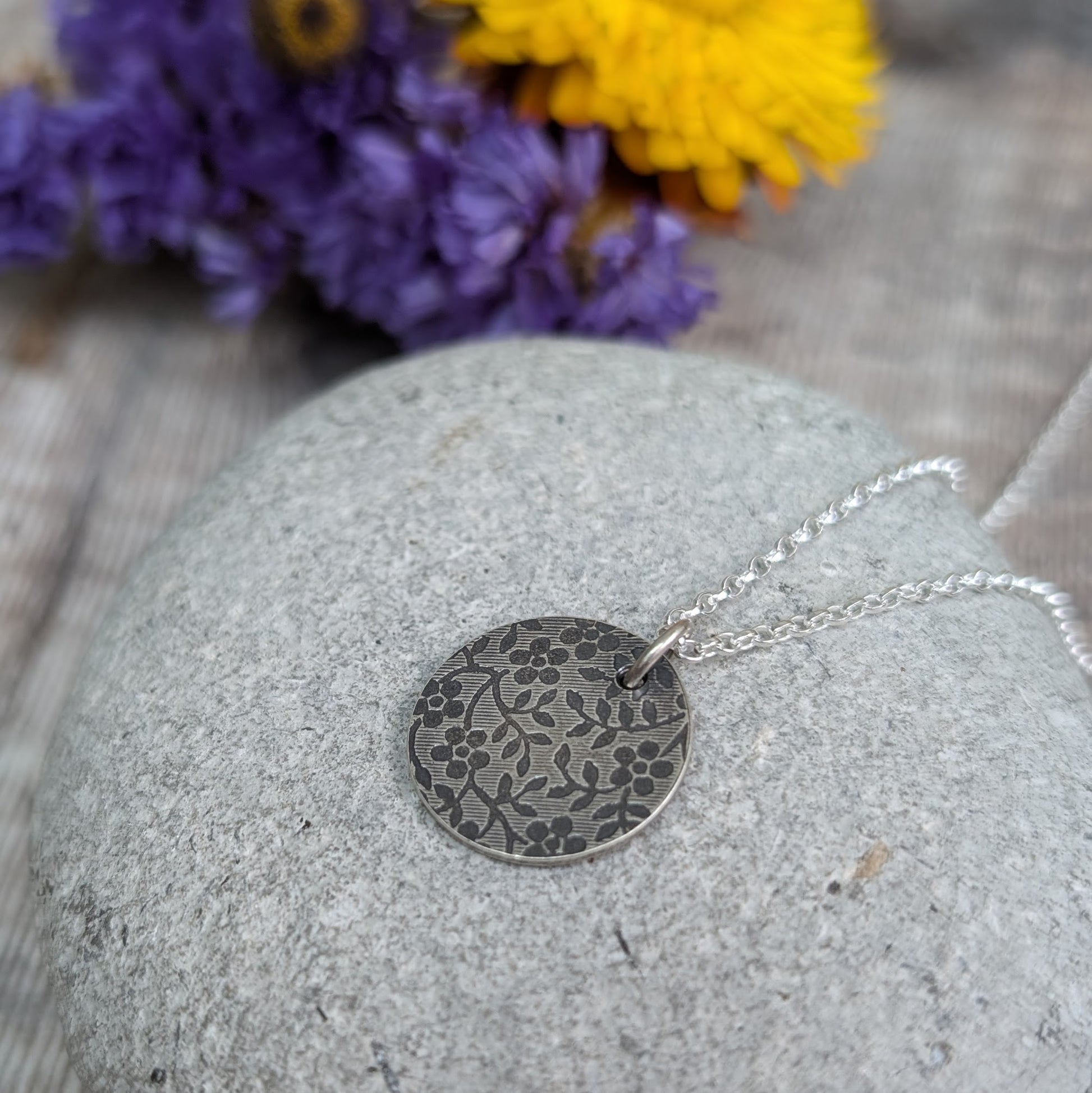 Pendant is a Sterling Silver oxidised disc stamped with a floral random pattern of five petalled flowers and small leaves on branches. Attached to a silver chain via small silver circle link.