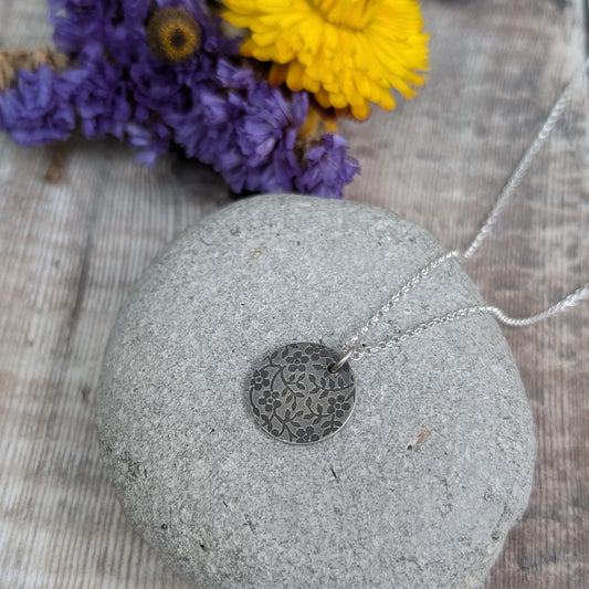 Pendant is a Sterling Silver oxidised disc stamped with a floral random pattern of five petalled flowers and small leaves on branches. Attached to a silver chain via small silver circle link.