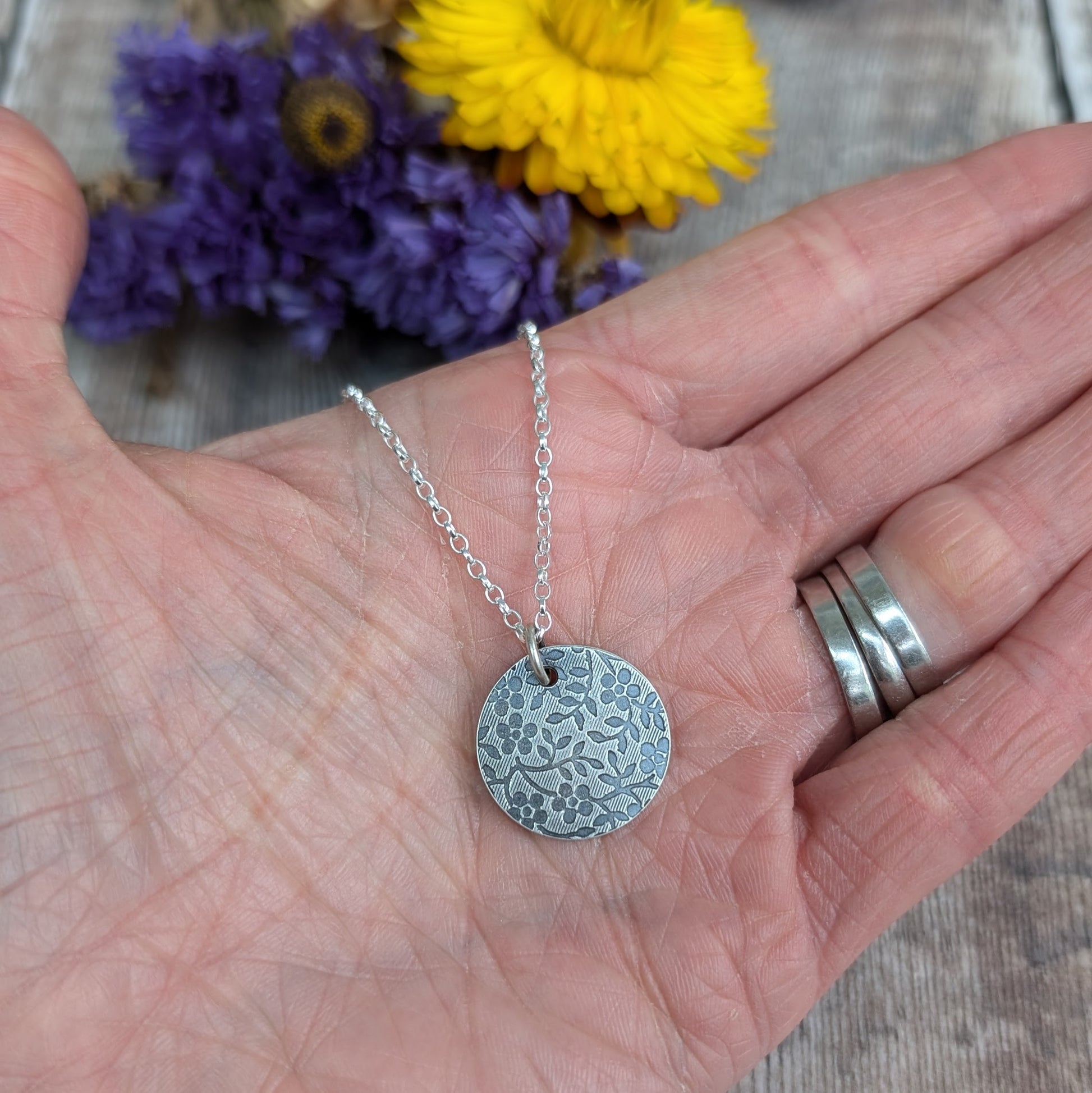Pendant is a Sterling Silver oxidised disc stamped with a floral random pattern of five petalled flowers and small leaves on branches. Attached to a silver chain via small silver circle link.