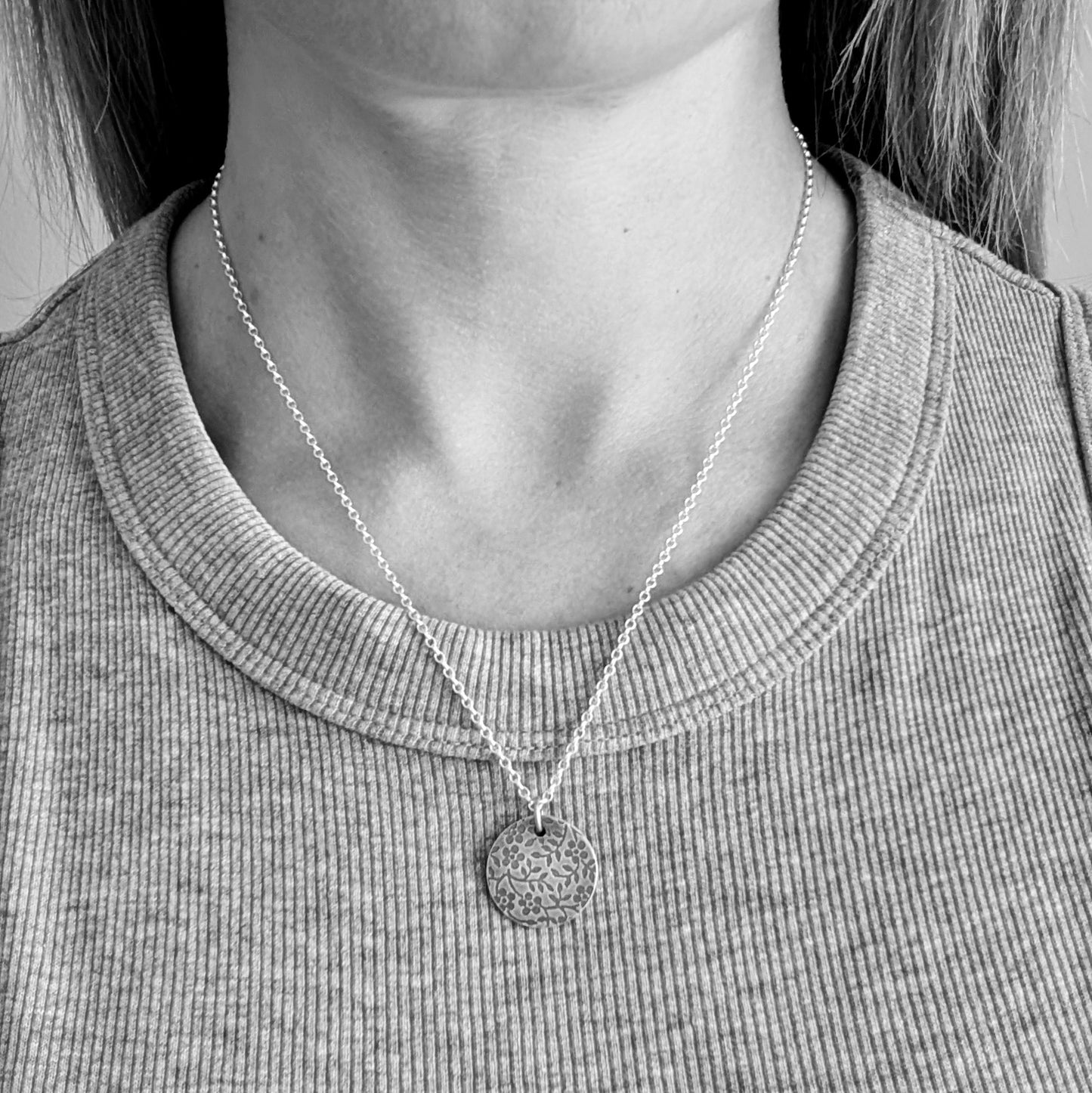 Pendant is a Sterling Silver oxidised disc stamped with a floral random pattern of five petalled flowers and small leaves on branches. Attached to a silver chain via small silver circle link.