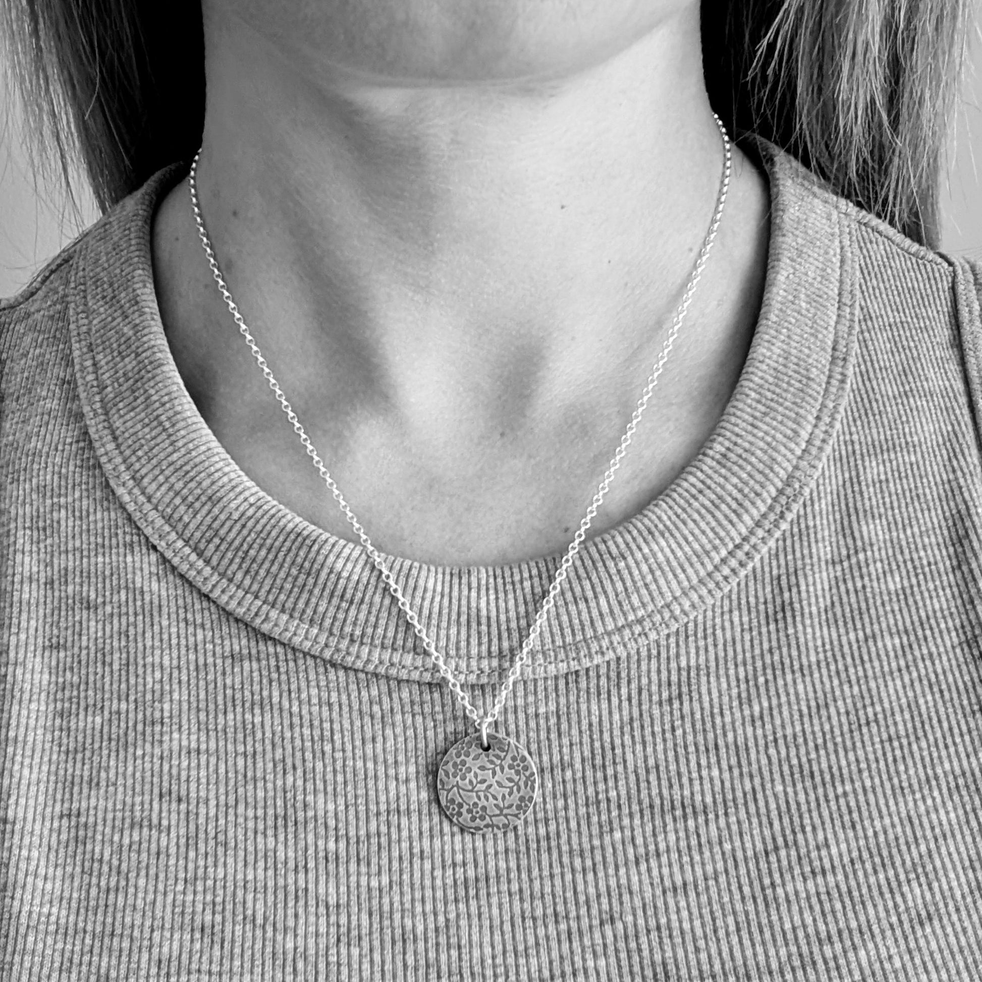 Pendant is a Sterling Silver oxidised disc stamped with a floral random pattern of five petalled flowers and small leaves on branches. Attached to a silver chain via small silver circle link.