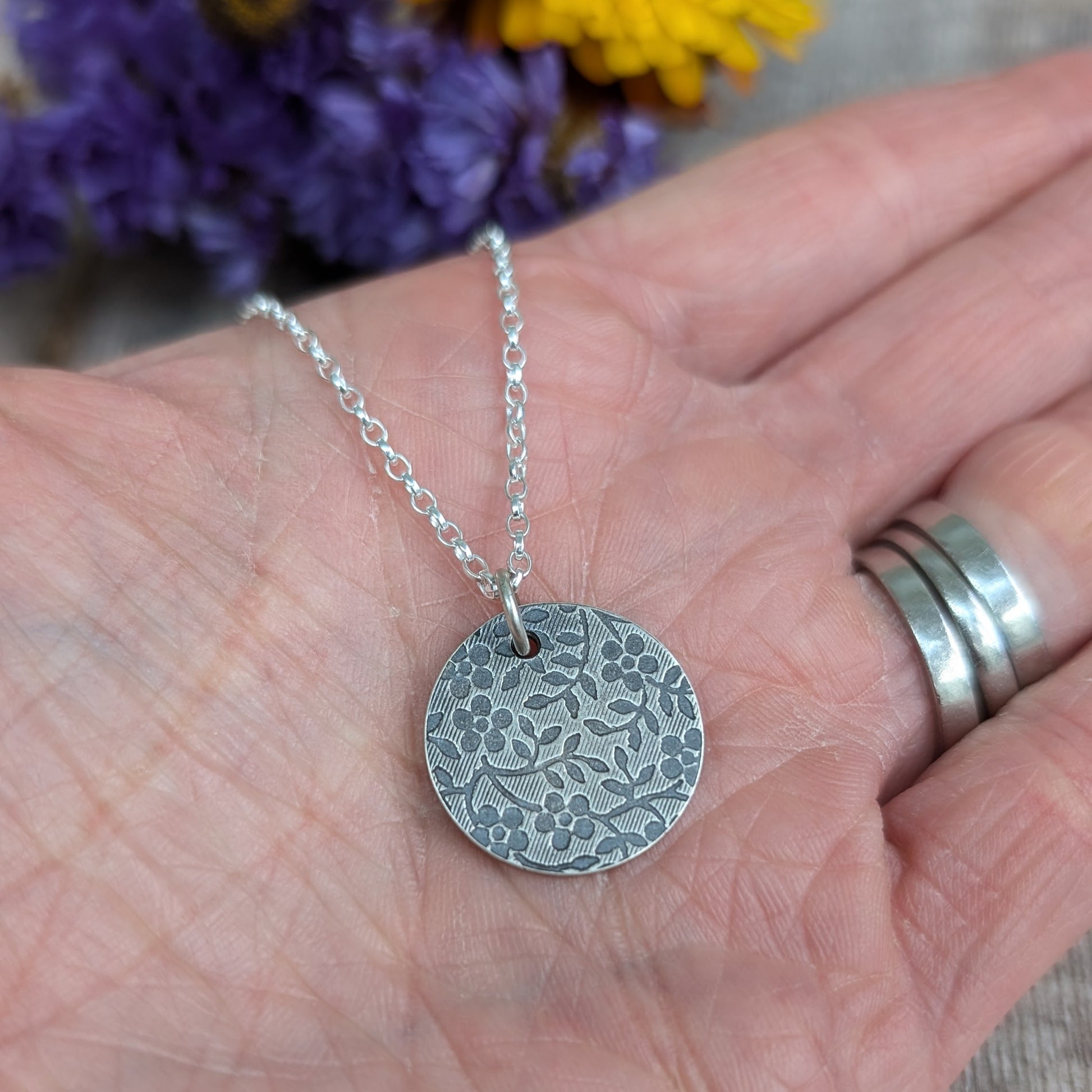 Pendant is a Sterling Silver oxidised disc stamped with a floral random pattern of five petalled flowers and small leaves on branches. Attached to a silver chain via small silver circle link.