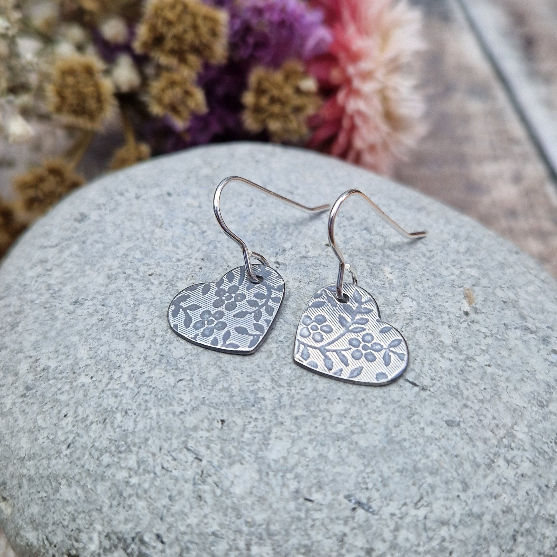 Each earring is an oxidised Sterling Silver heart shape hanging from silver earring wire. The heart shape is stamped with a floral pattern of flowers with 5 petals and leaves.