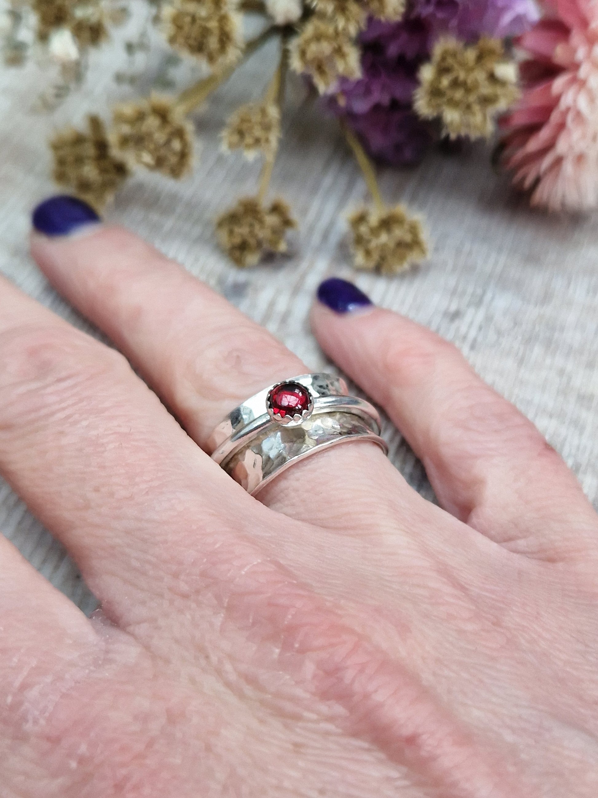 Approx. 10mm wide hammered texture silver band with additional 2mm wide silver band around the middle that spins independently. Mounted in a silver bezel on top of the 2mm wide band is a round red garnet gemstone, approx. 5mm diameter.