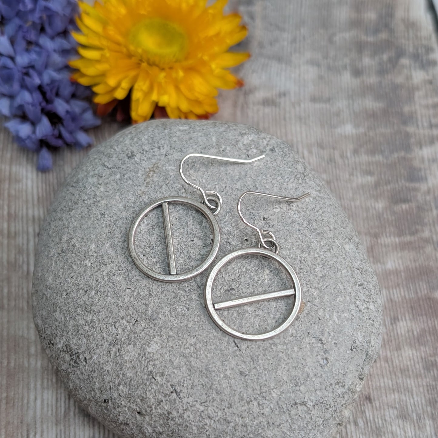 A pair of handmade silver earrings with open circular frames and a diagonal bar across each, resting on a smooth grey stone. A yellow flower is in the background, slightly out of focus.