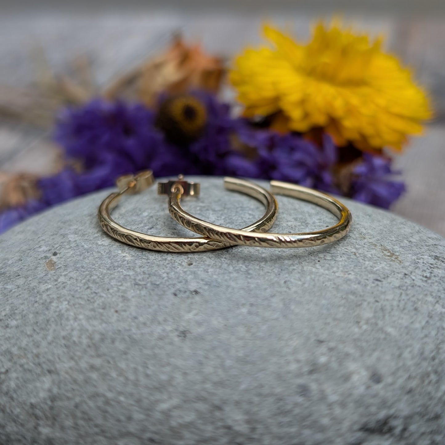 Pair of gold hoop earrings featuring the 'Meadow' pattern, displayed on a smooth stone with purple and yellow flowers in the background, highlighting the handcrafted details and recycled 9ct gold material.