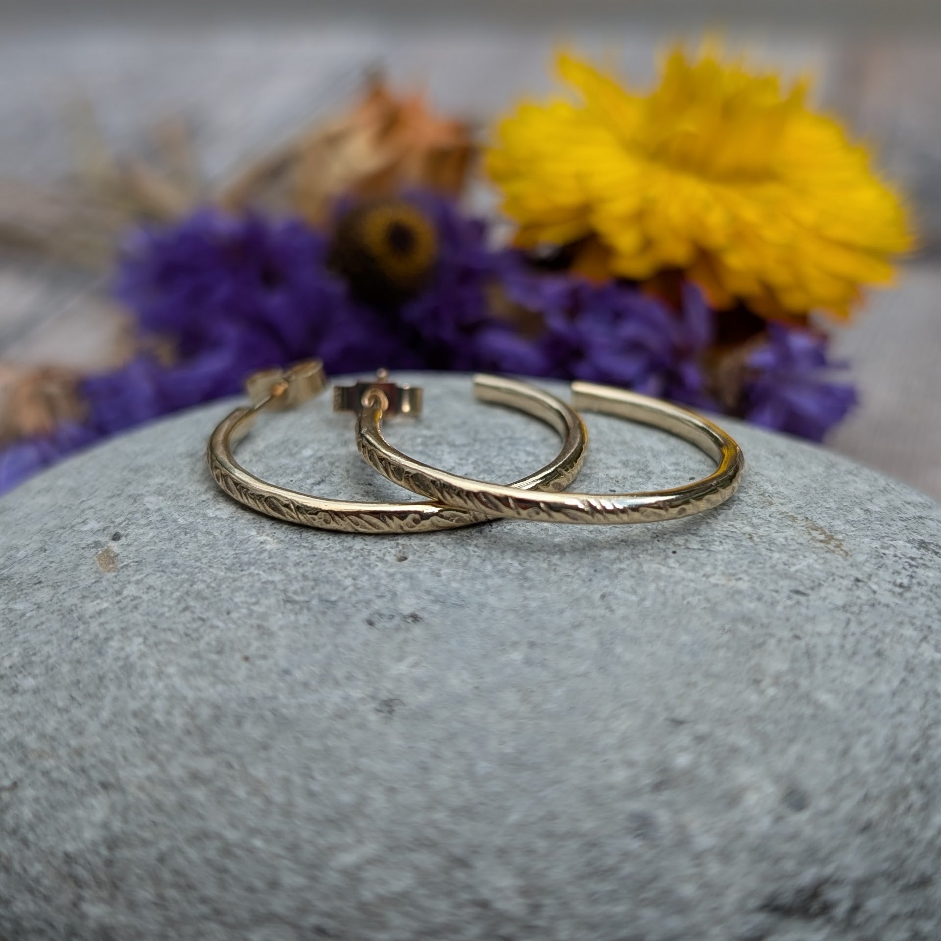 Pair of gold hoop earrings featuring the 'Meadow' pattern, displayed on a smooth stone with purple and yellow flowers in the background, highlighting the handcrafted details and recycled 9ct gold material.