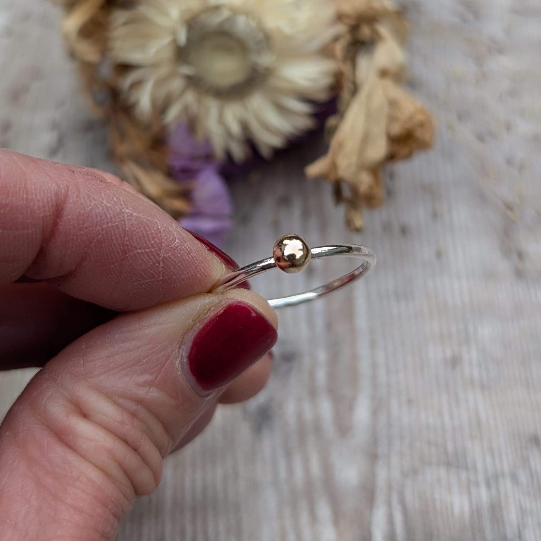 A hand holding a recycled silver ring with a small gold pebble, displaying the ring’s minimalist design against a neutral background with dried flowers.