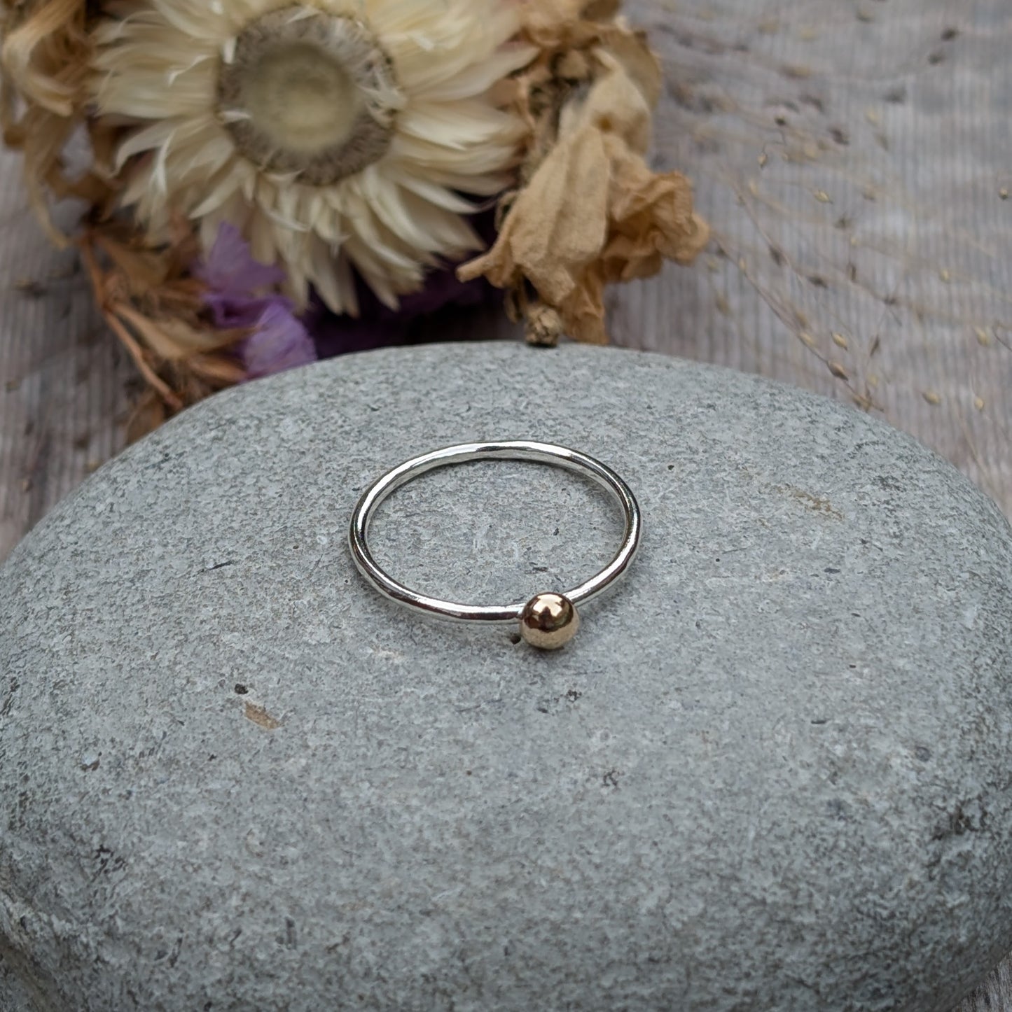 A close-up of a recycled silver ring with a gold pebble, placed on a grey stone surface with dried flowers in the background, adding a natural and earthy vibe to the presentation.