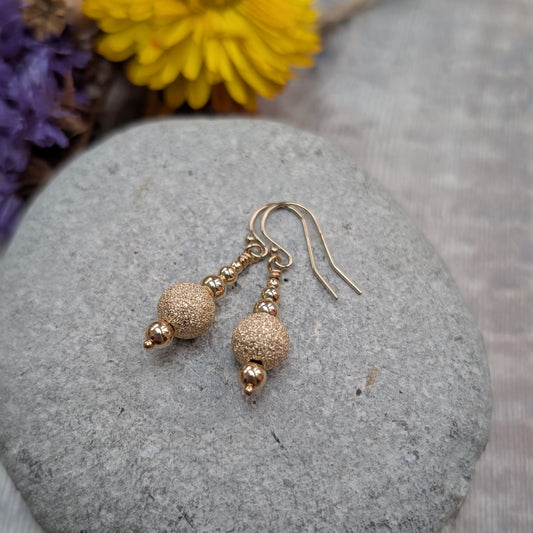 Detailed view of gold filled earrings with textured gold sparkle beads and polished gold beads, styled on a grey stone with soft, natural props.