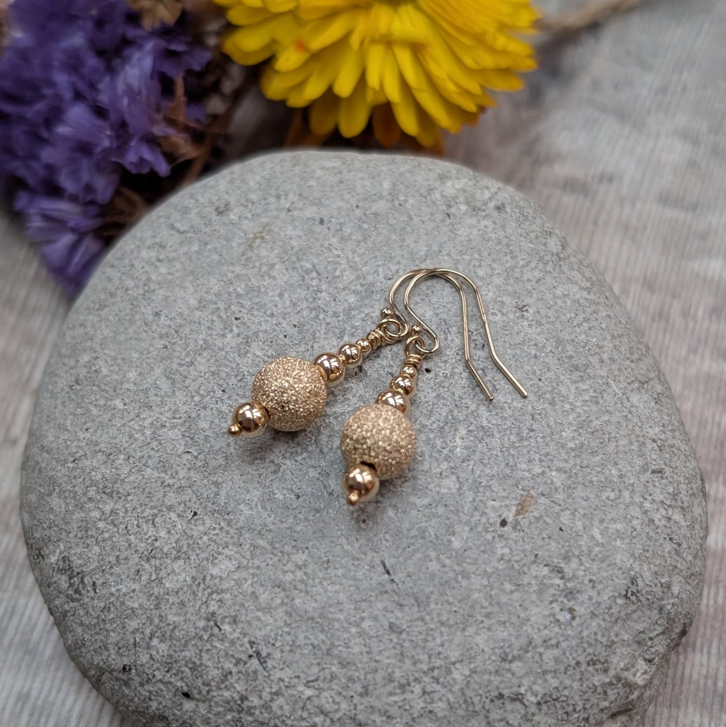Pair of gold filled earrings with textured gold sparkle beads, displayed on a smooth grey stone with dried flowers in the background.
