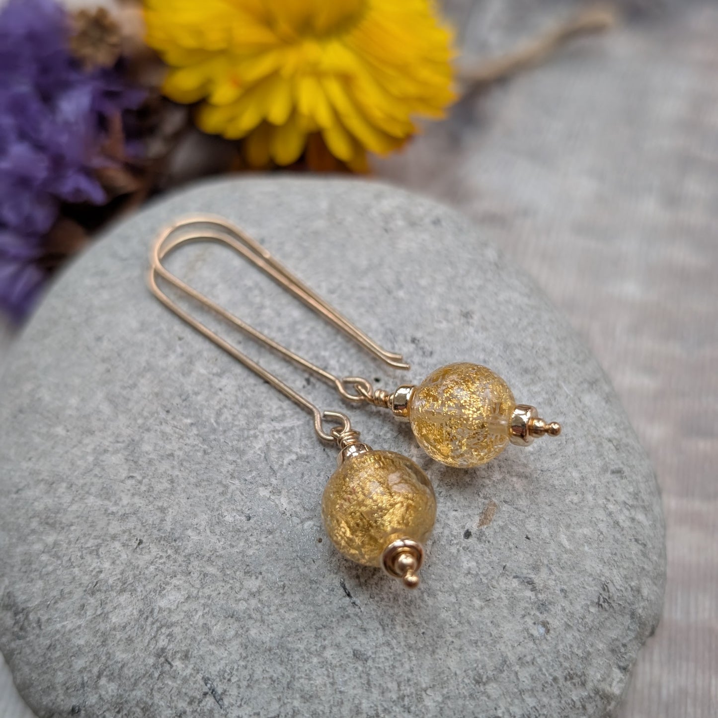 Pair of gold filled long earrings with gold fleck Venetian glass beads, displayed on a grey stone with soft, natural styling.